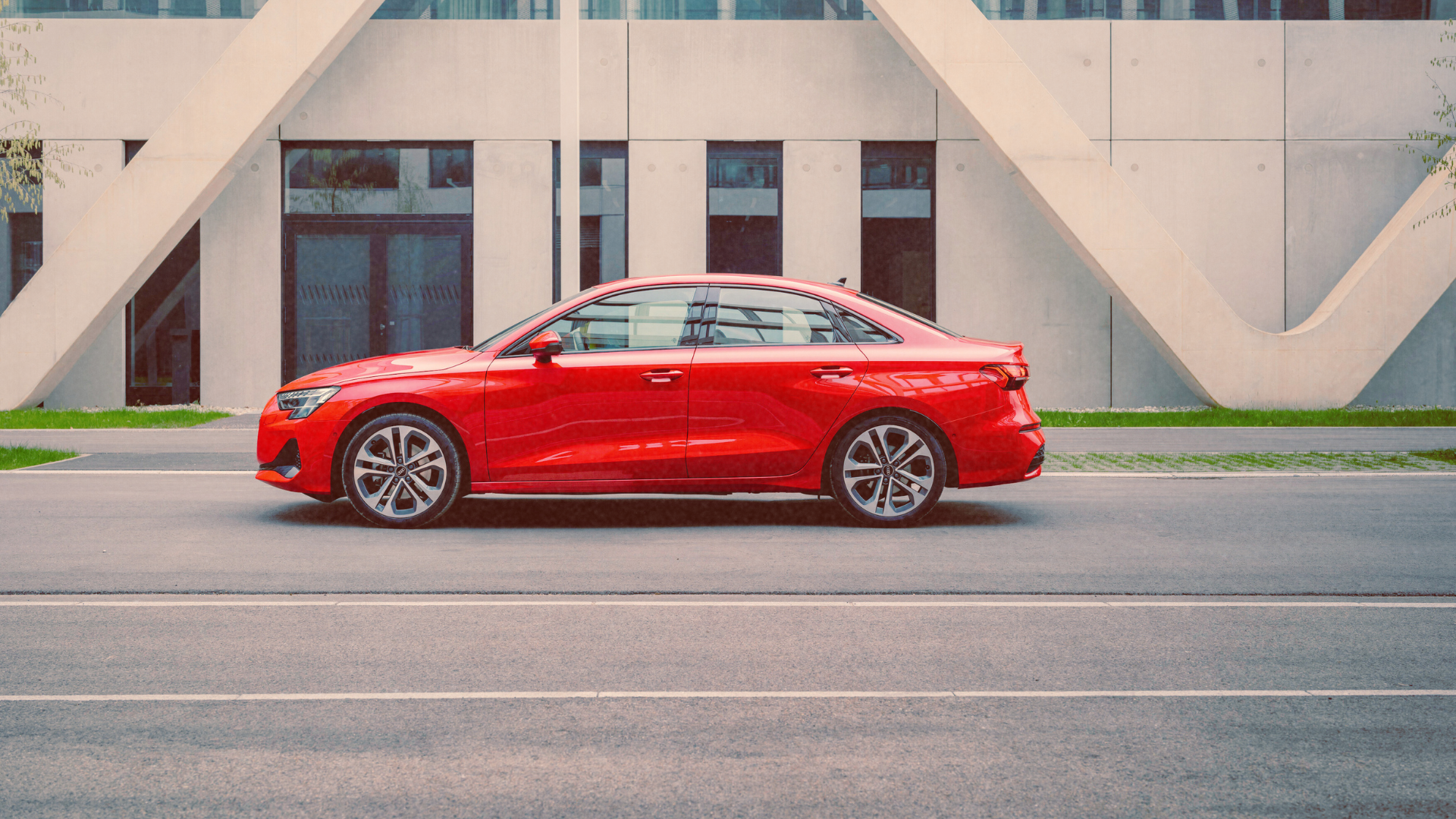 Side profile of an Audi A3 red luxury sedan parked on a city street in front of modern architecture.