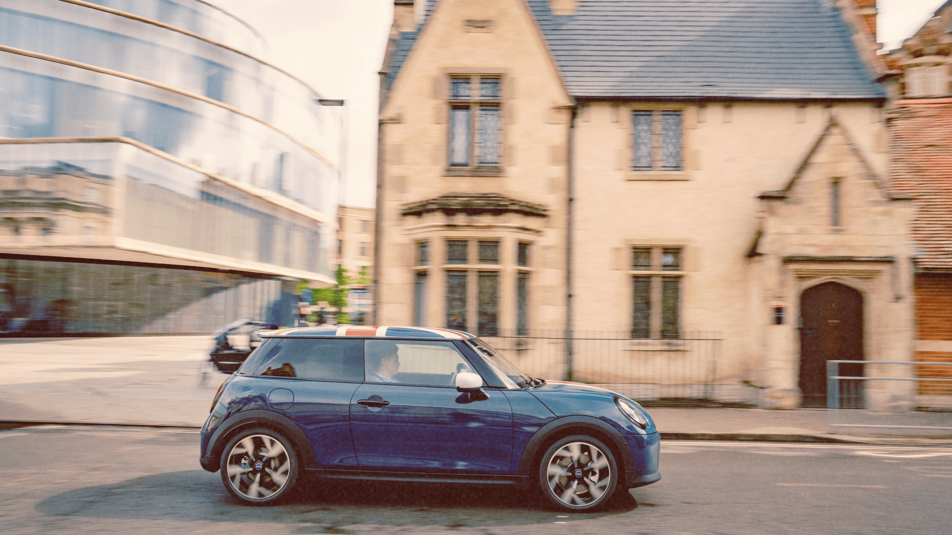 A blue Mini Cooper with a Union Jack roof driving past historic and modern buildings in Oxford.