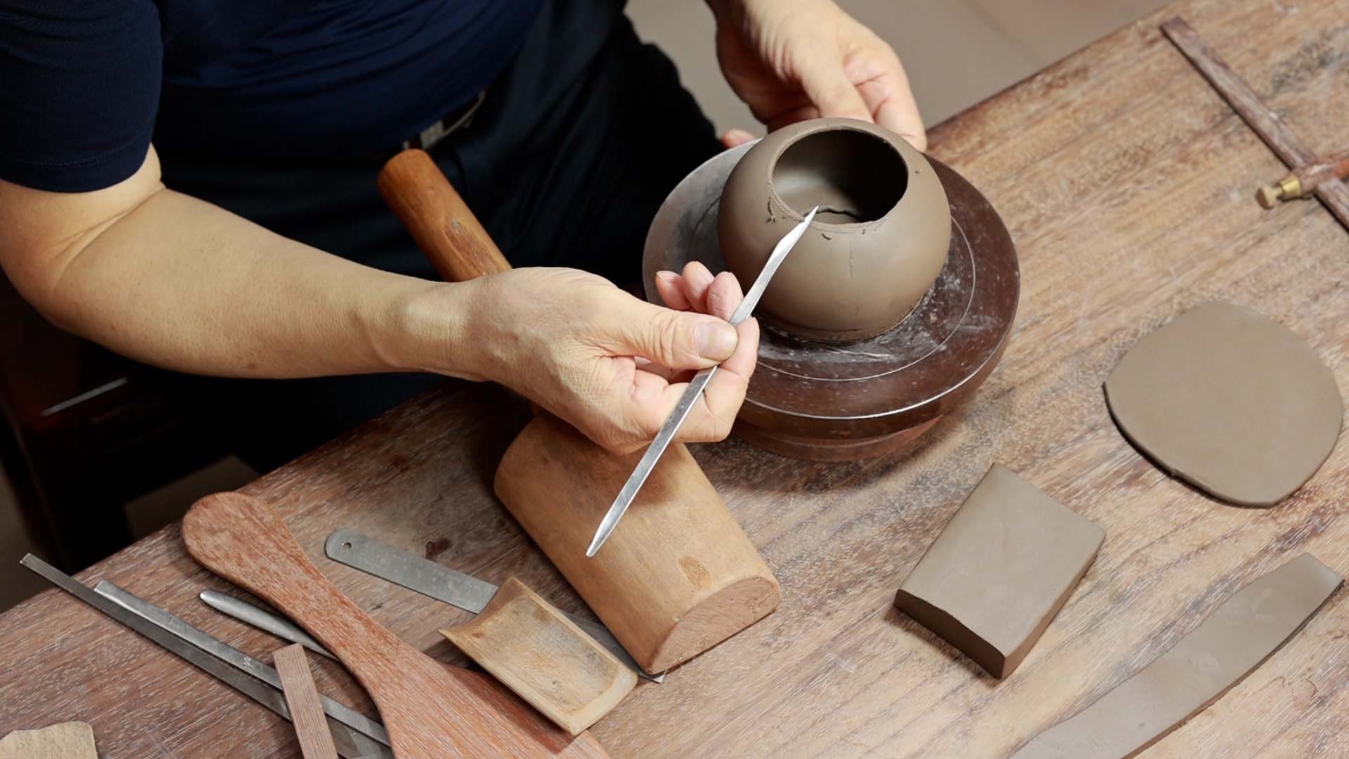 A man holding a long-handled pointed knife, carving the edge of a Yixing teapot