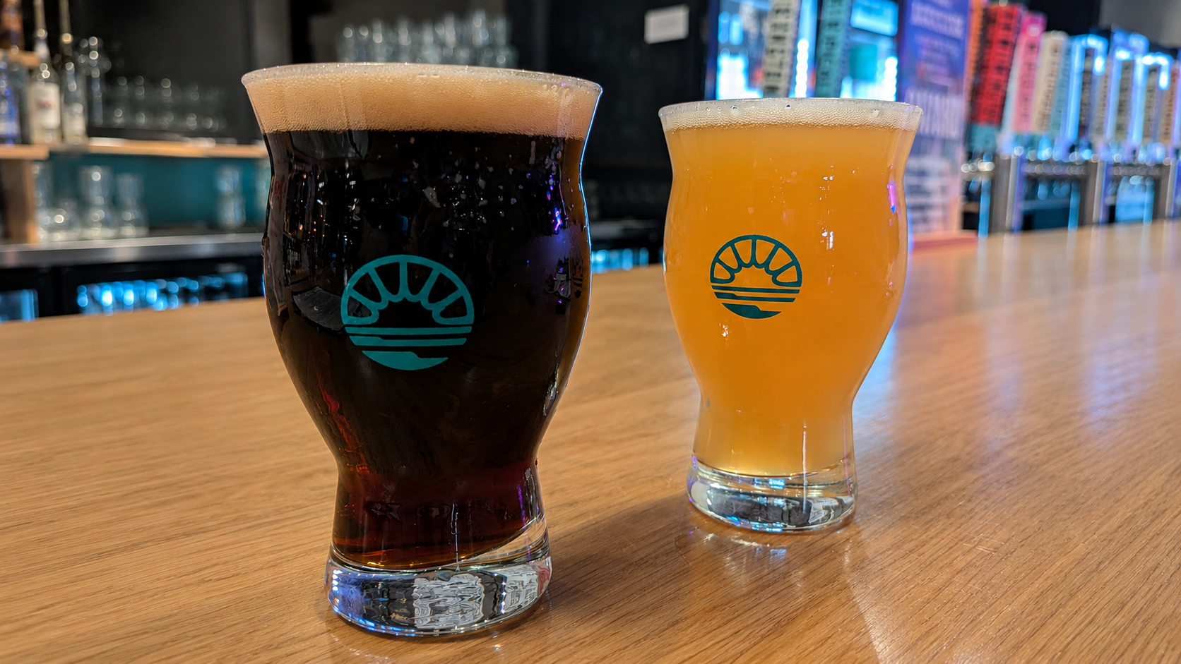 A dark stout and a hazy IPA craft beer served in tulip glasses on a wooden bar counter.