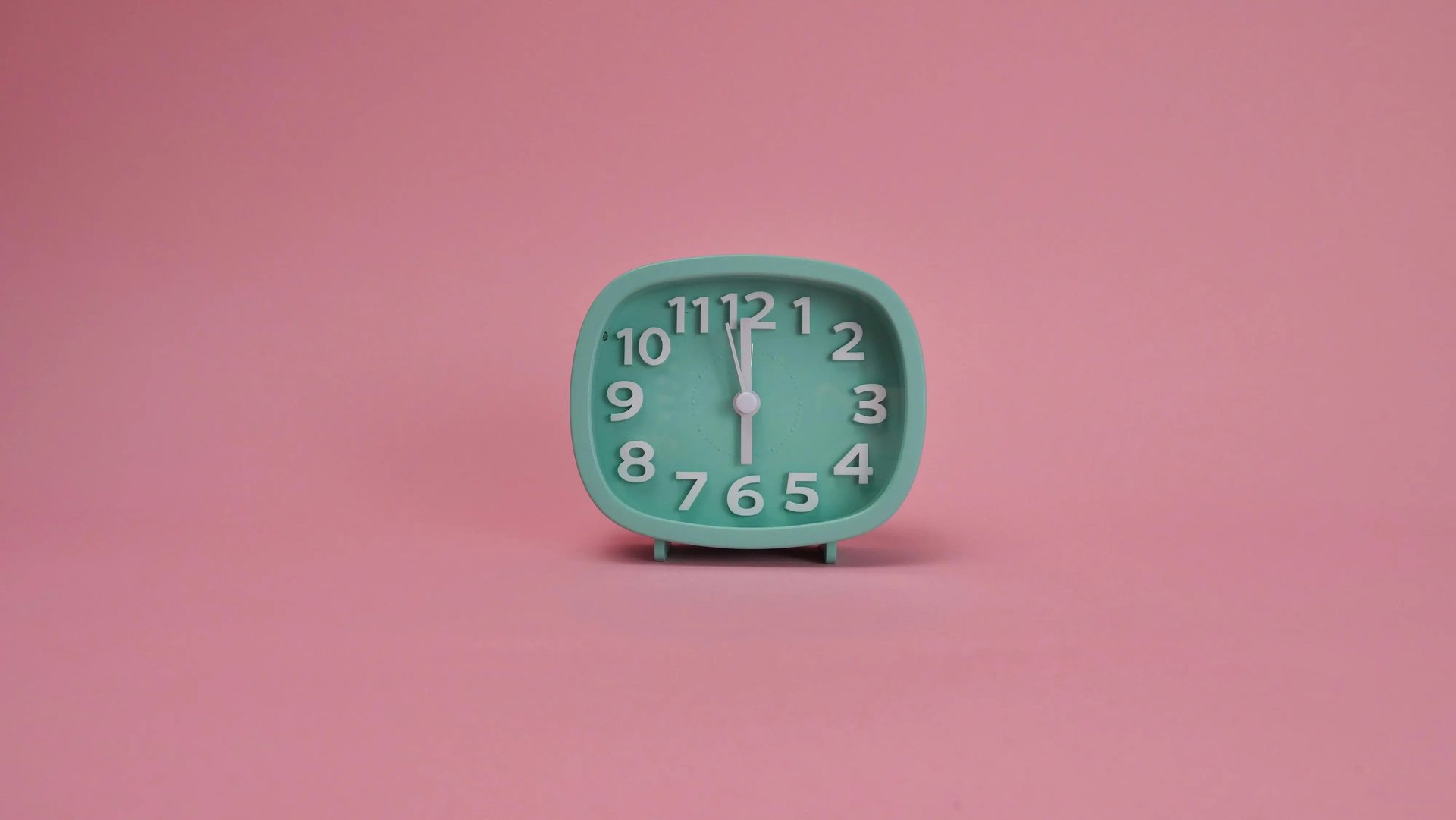 A small mint green analog alarm clock showing six o'clock against a solid pink background.