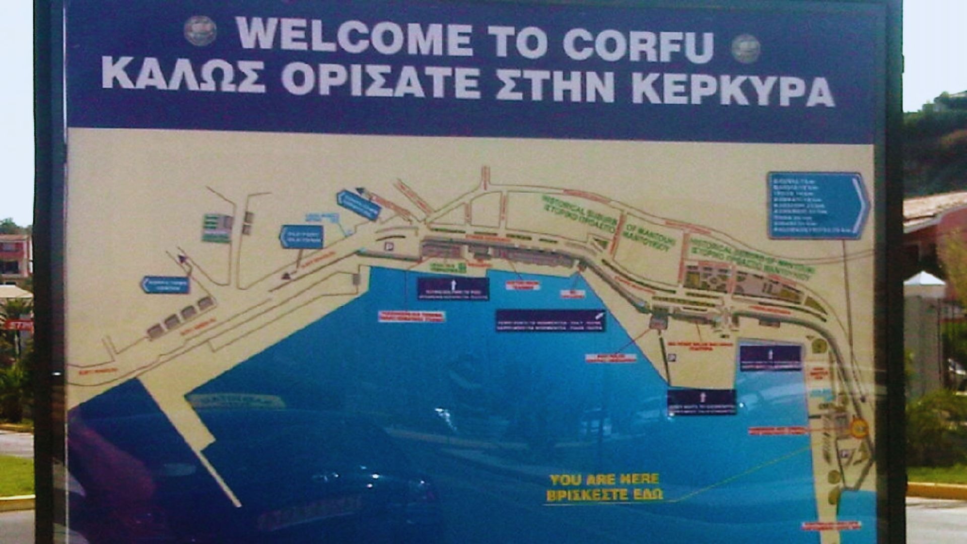 Sinal de boas-vindas no porto de Corfu com vista para o mar e estrutura portuária ao fundo.