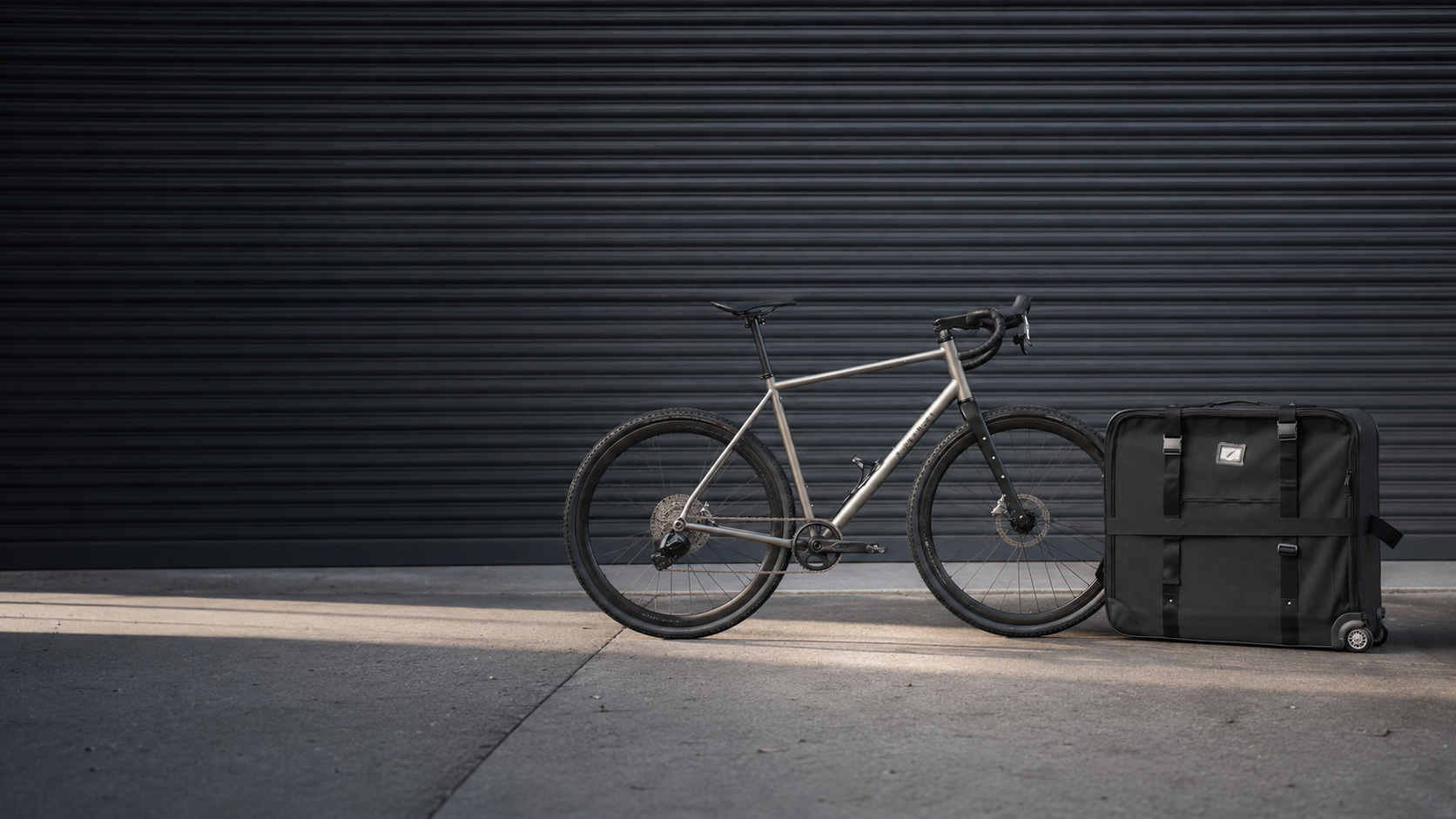 A titanium gravel bike leaning against a black travel bike bag in front of a dark shutter door.