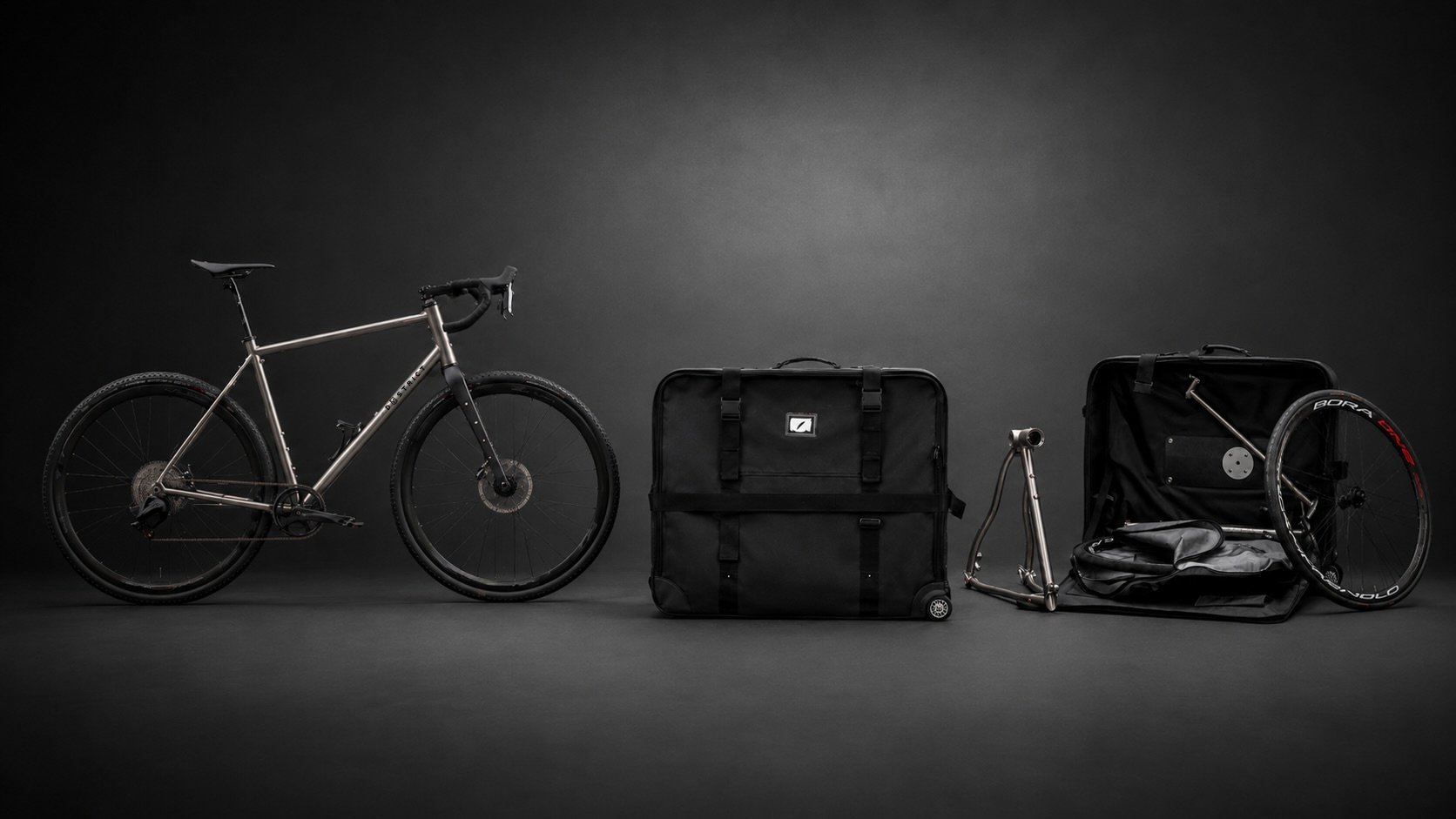 Titanium gravel bike next to a black padded bike travel bag and open storage case.