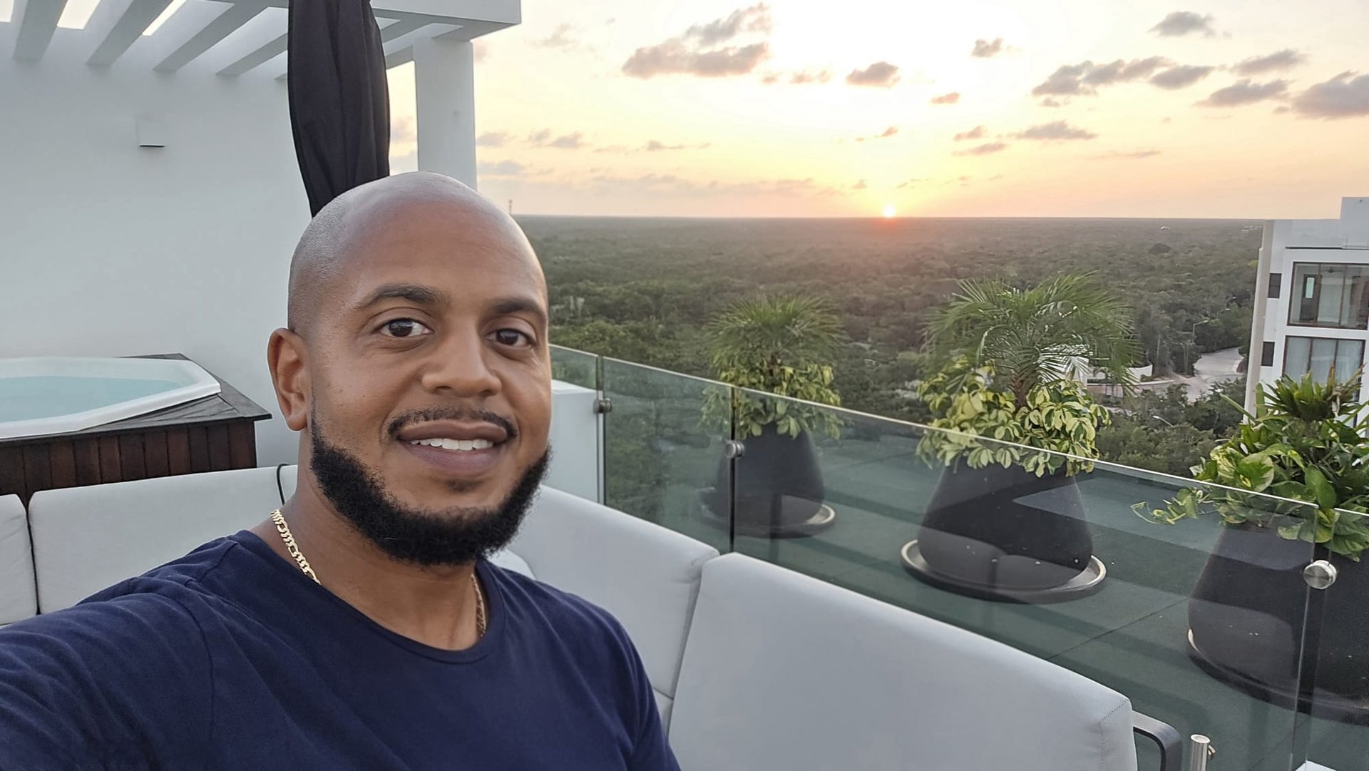 Smiling man taking a selfie on a luxury rooftop lounge at sunset overlooking a tropical forest.