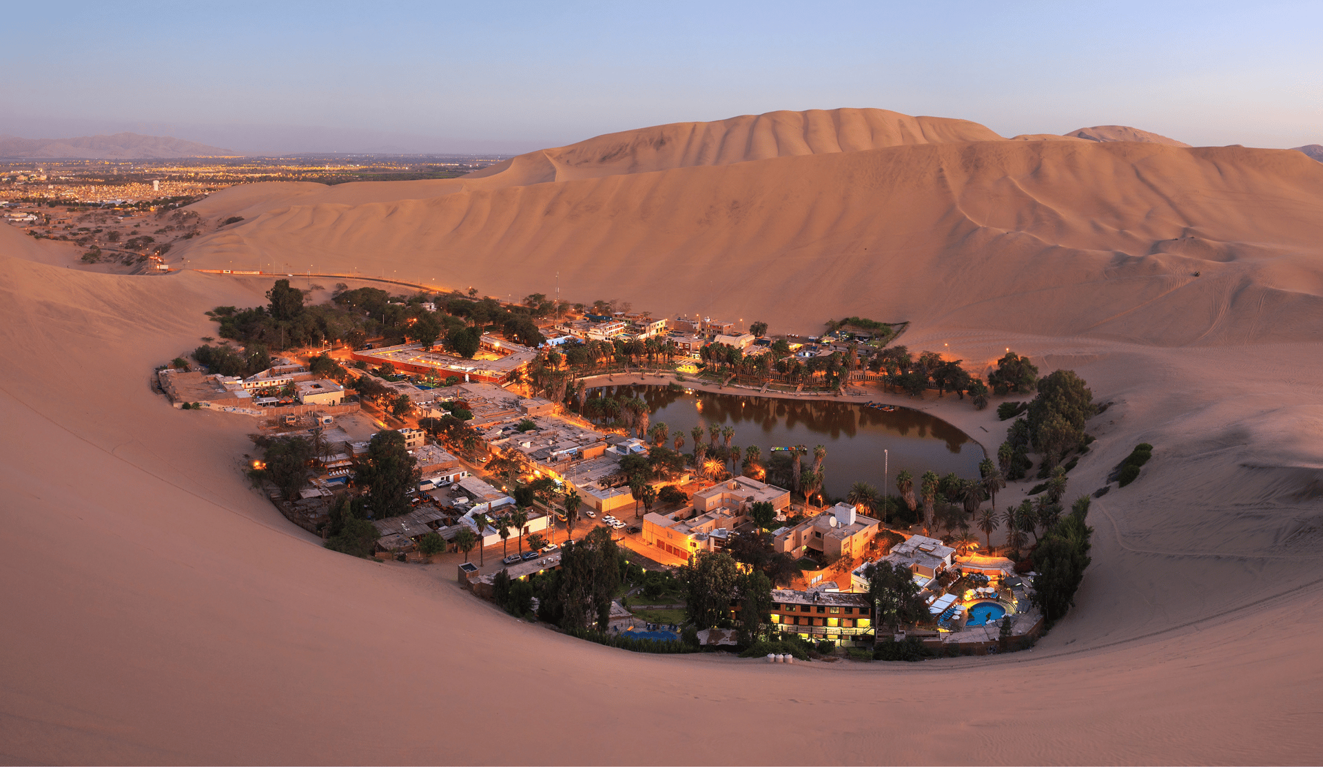 Aerial view of Huacachina desert oasis in Peru with illuminated village buildings and palm trees at dusk.