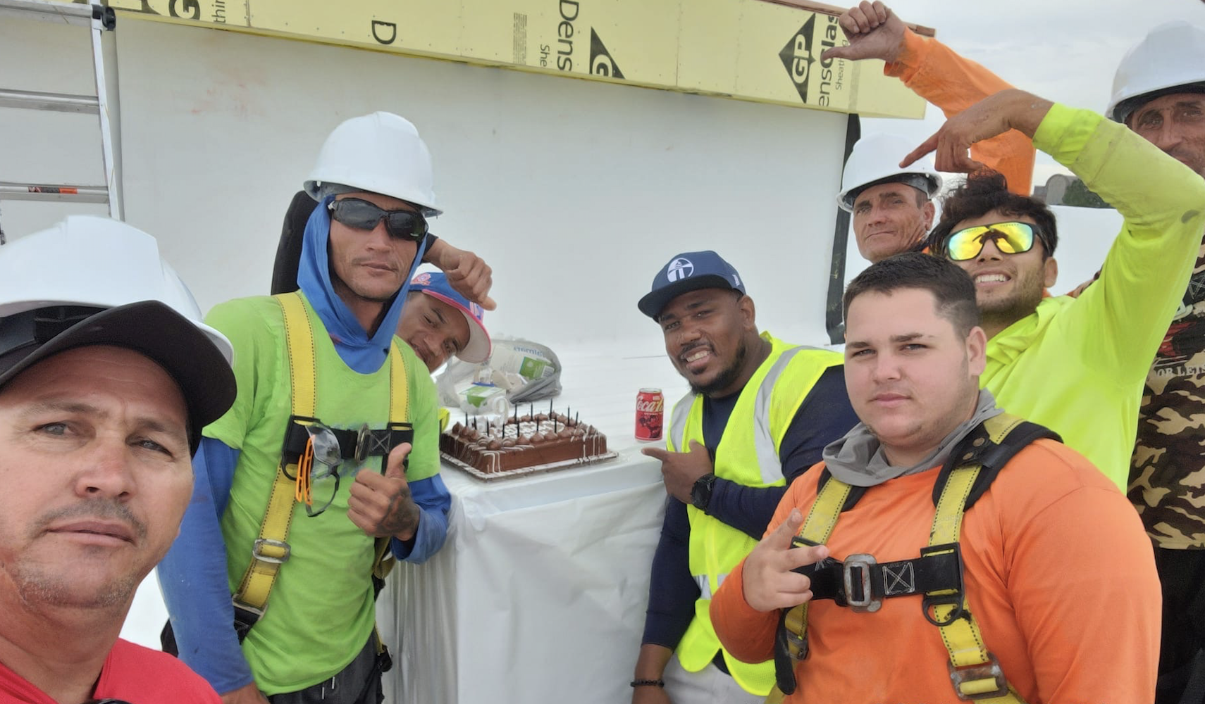 Construction workers in hard hats and safety vests celebrating with a chocolate cake on a job site.