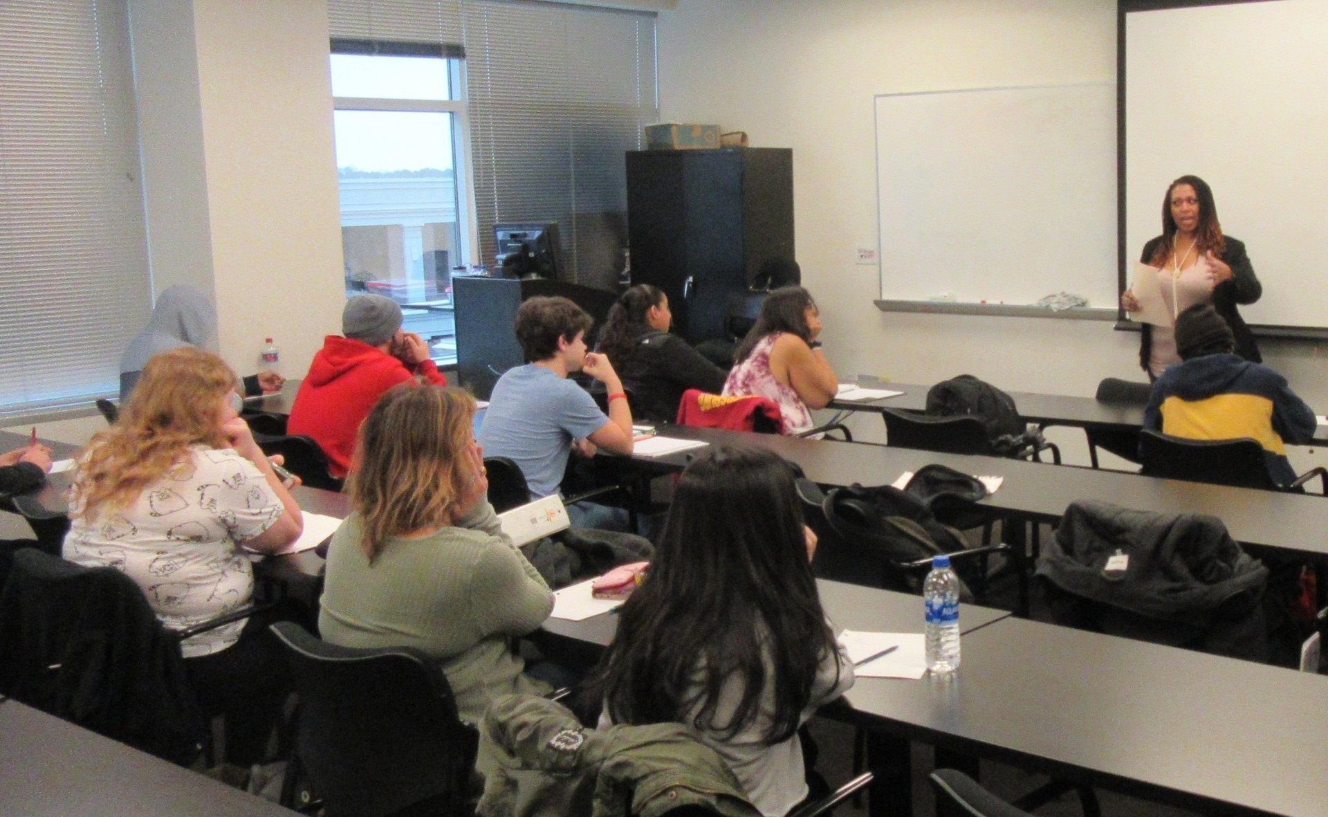 A diverse group of students in a classroom listening to a female teacher giving a lecture.
