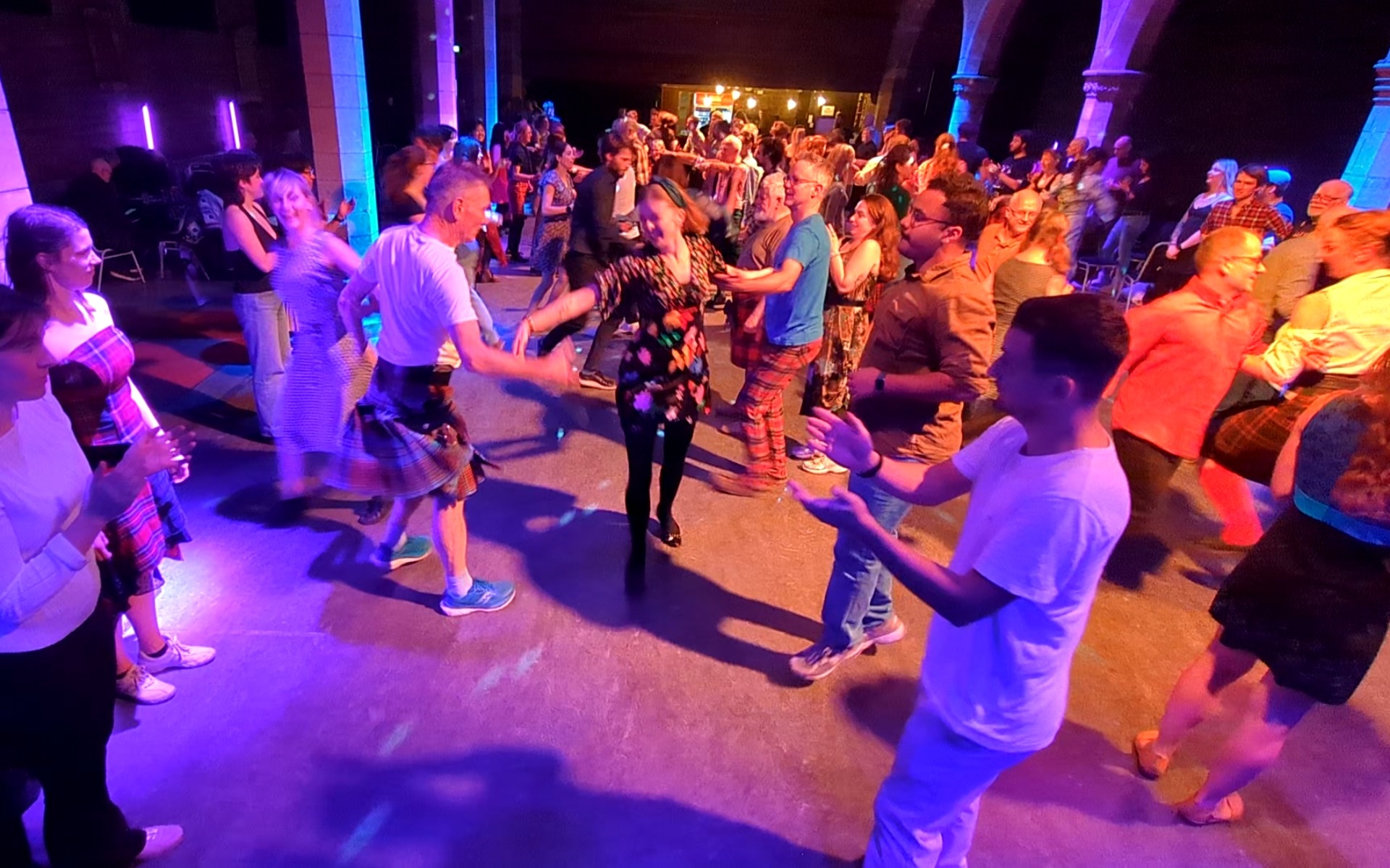 People dancing at a traditional Scottish ceilidh night with a live band and purple stage lighting.