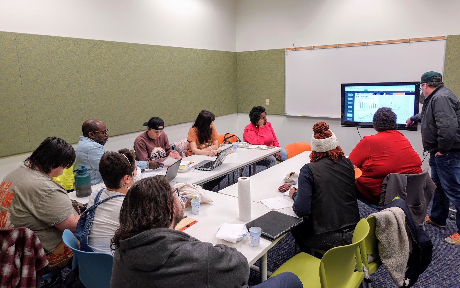 A group of people gathered around a table, looking intently at a presenter in the front of the room