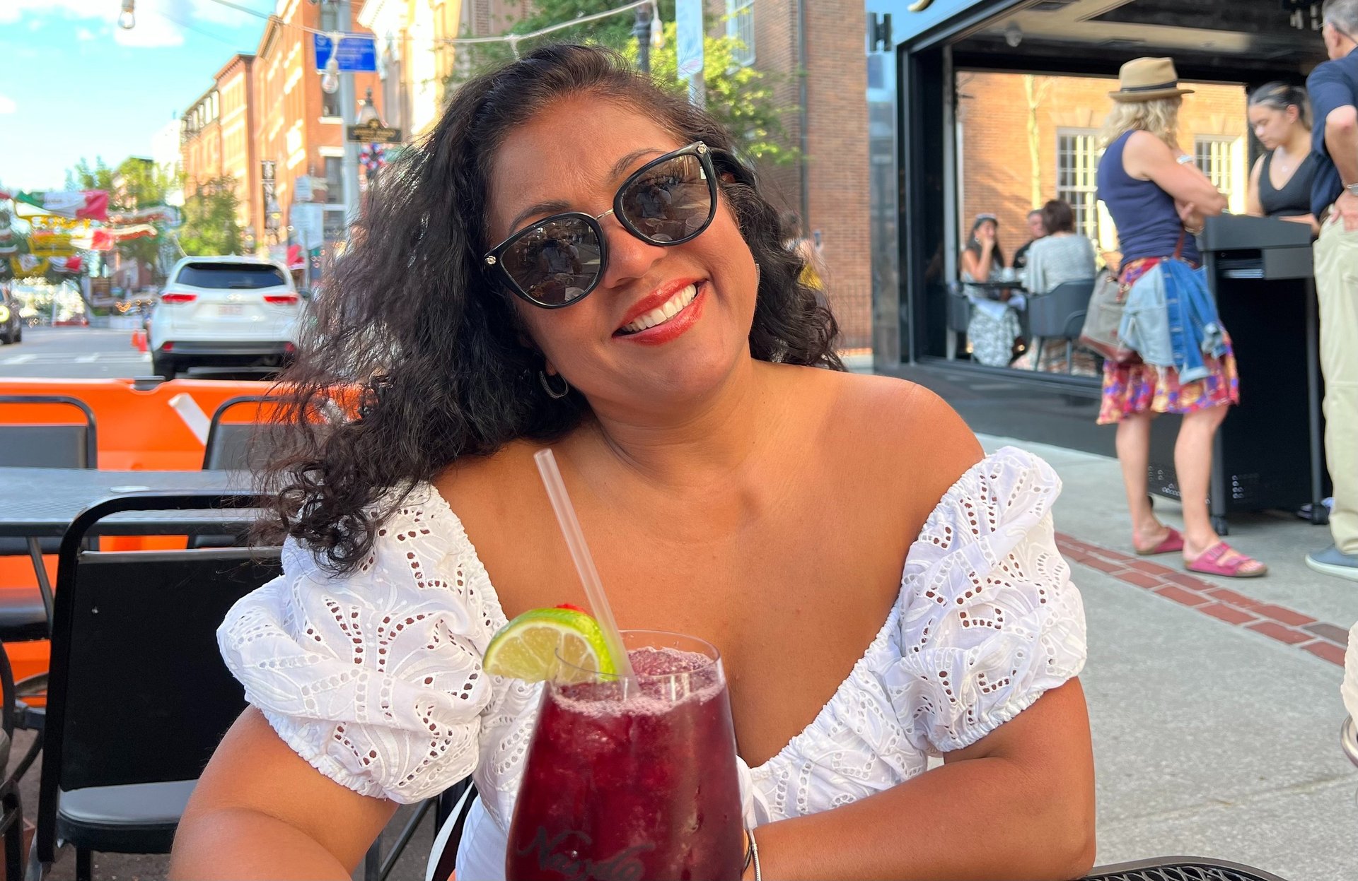 Smiling woman in a white eyelet top enjoying a red sangria cocktail at an outdoor city patio