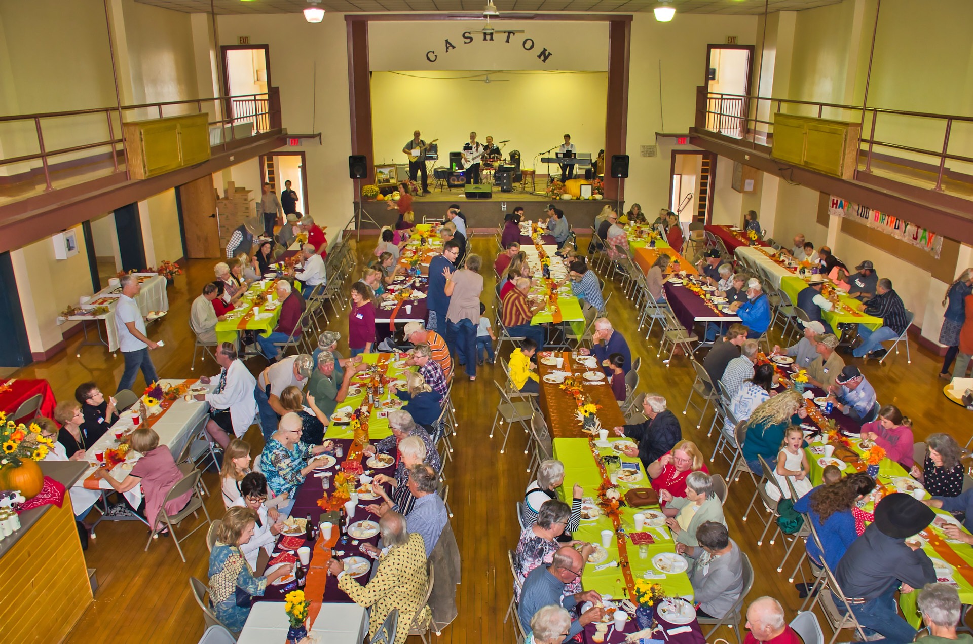 a large group of people sitting at tables with food eating together with a band on the stage