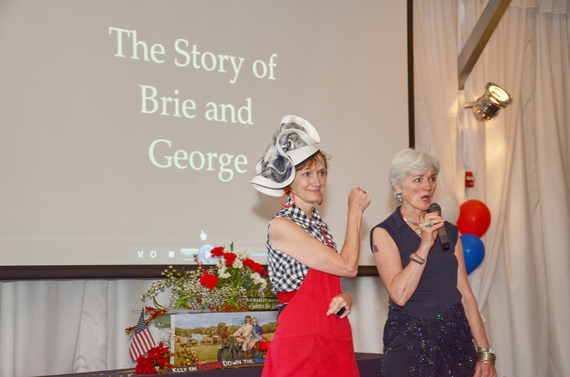 two women in a big hat and dresses standing in front of a screen speaking to the audience