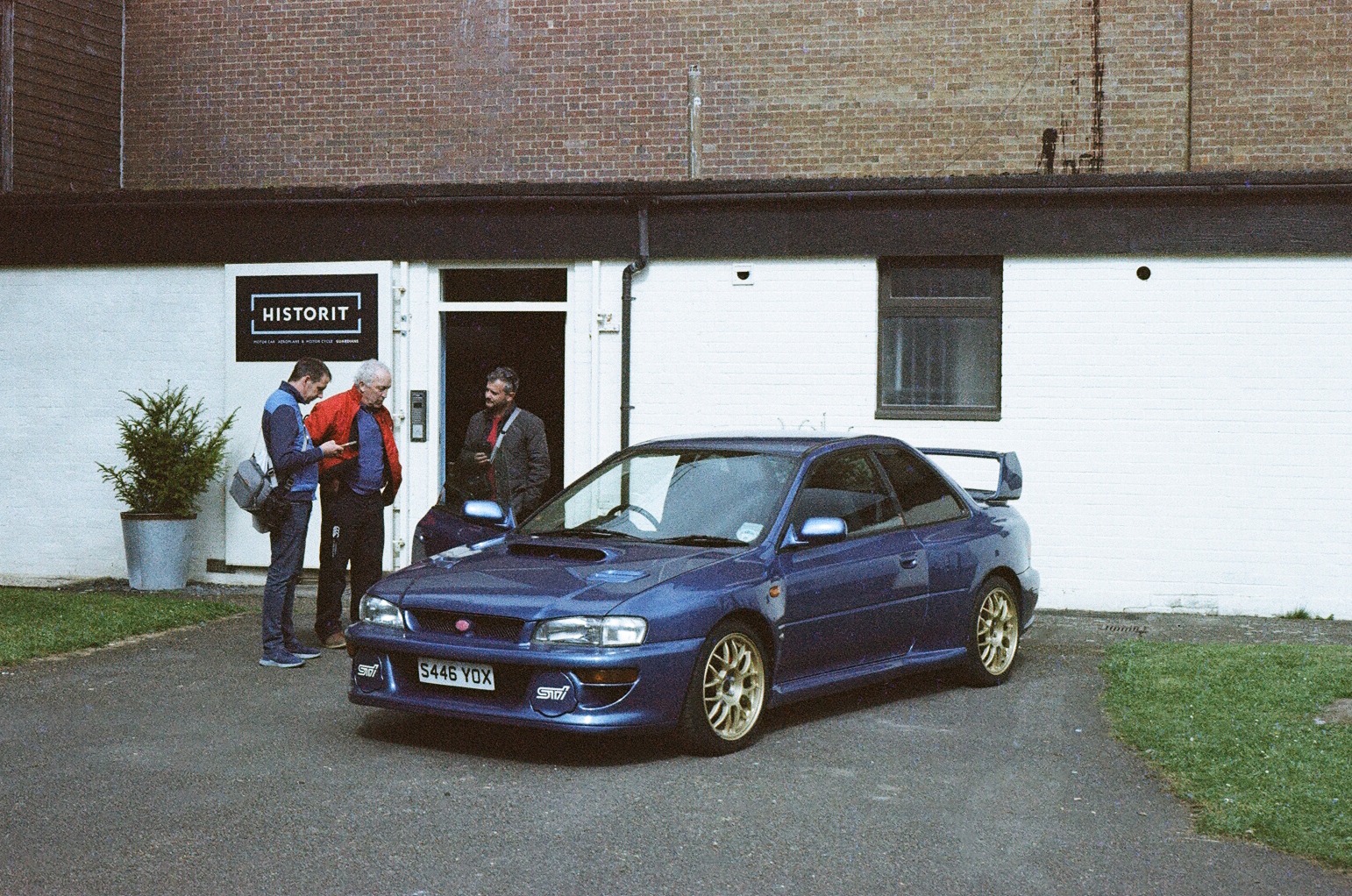 a classic Subaru Impreza parked in front a building with three guys looking at it