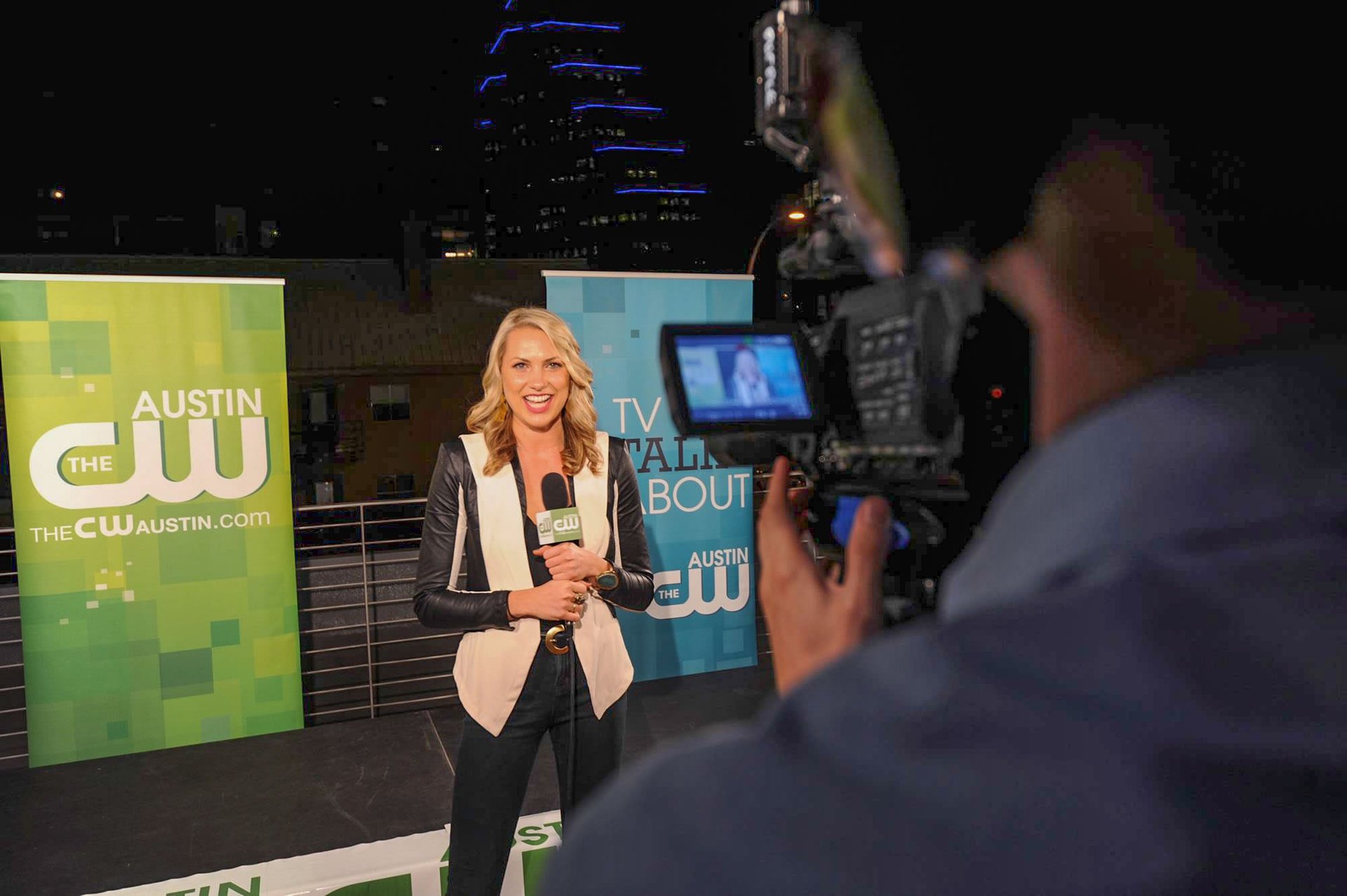 CW Star smiles on-camera, recording segment with pro gear against branded backdrop and city lights.