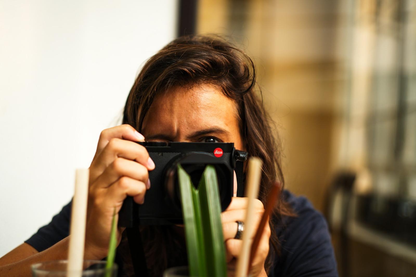 Close‑up of a person with a Leica camera, blurred green leaves in foreground - By ACAT Photos