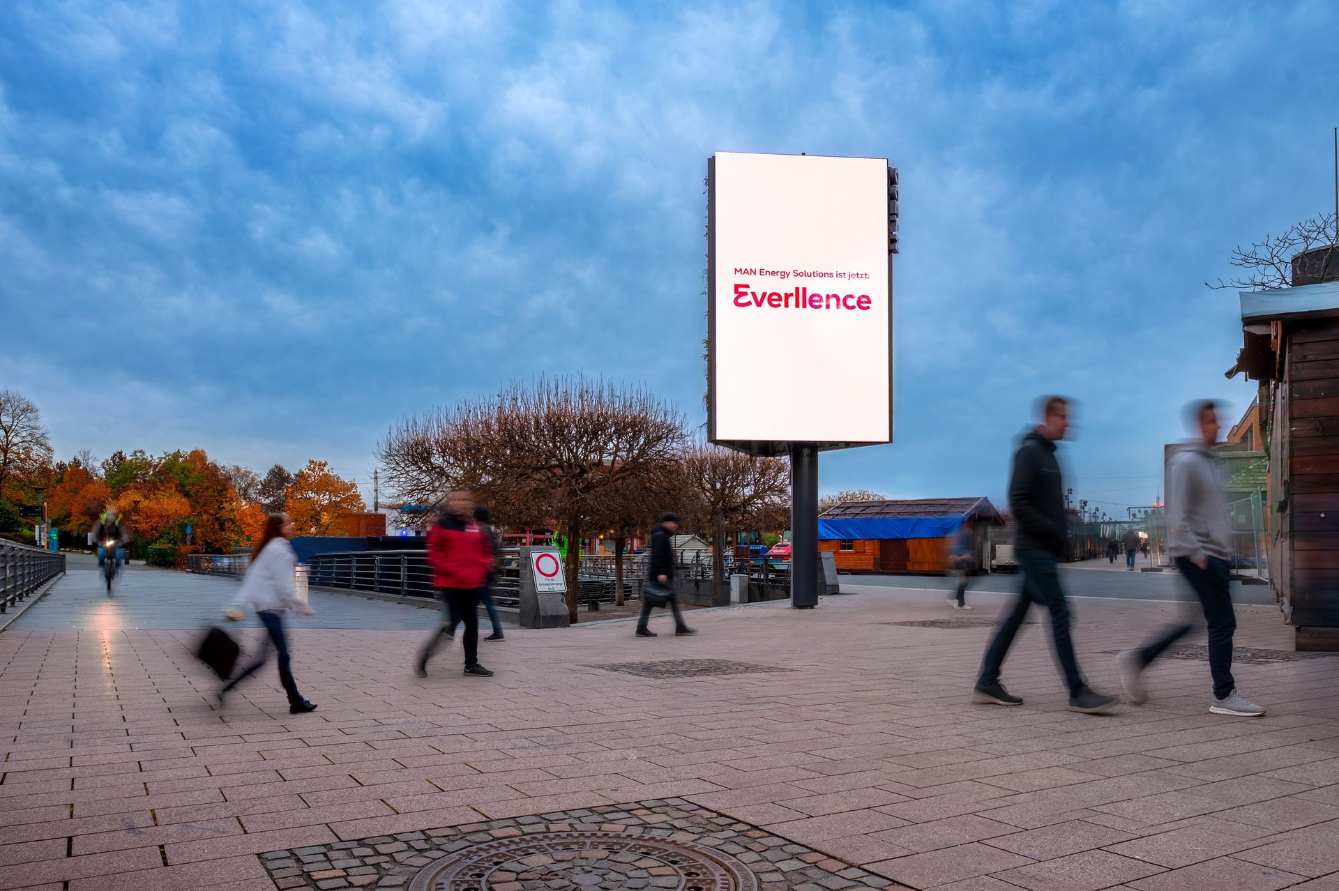 Large outdoor digital billboard displaying an Everllence advertisement in a busy pedestrian plaza with motion-blurred