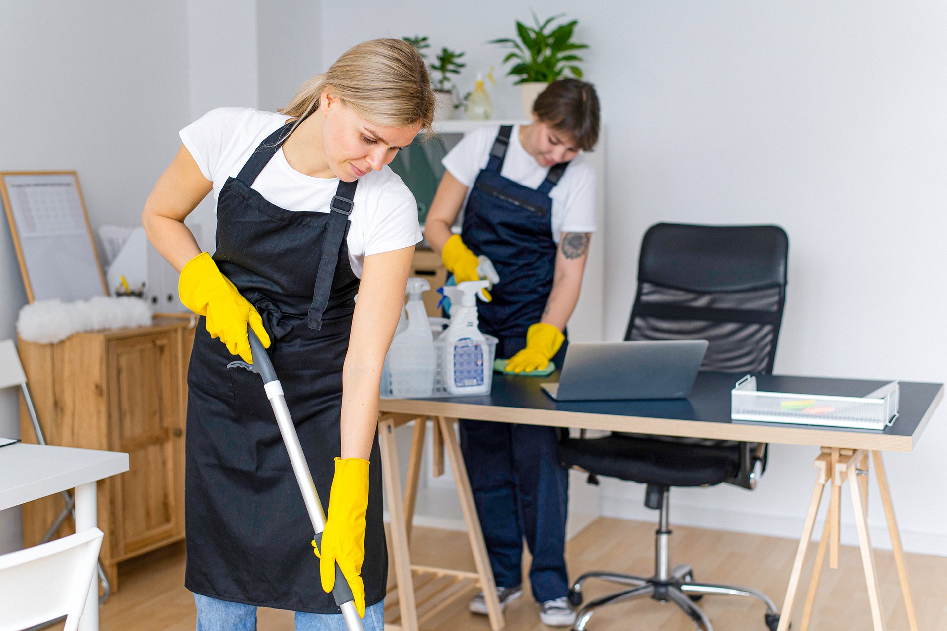 Dwie pracownice firmy sprzątającej w rękawiczkach czyszczą nowoczesne biuro – jedna mopuje podłogę, druga porządkuje biurko