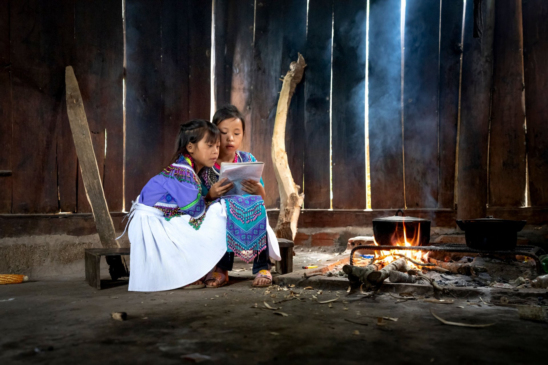 Two young girls in traditional attire reading together by an indoor cooking fire.
