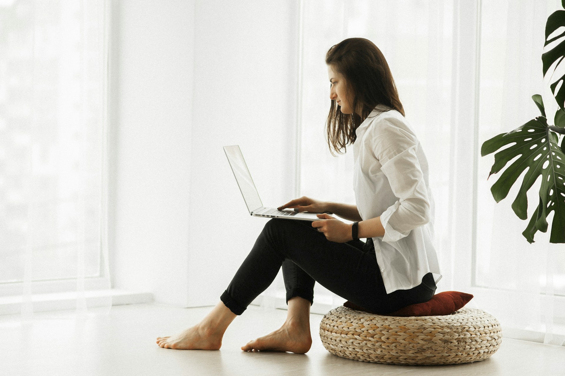 A woman in a white shire and black slacks, sitting on a floor cushion in a white room, working on a laptop.