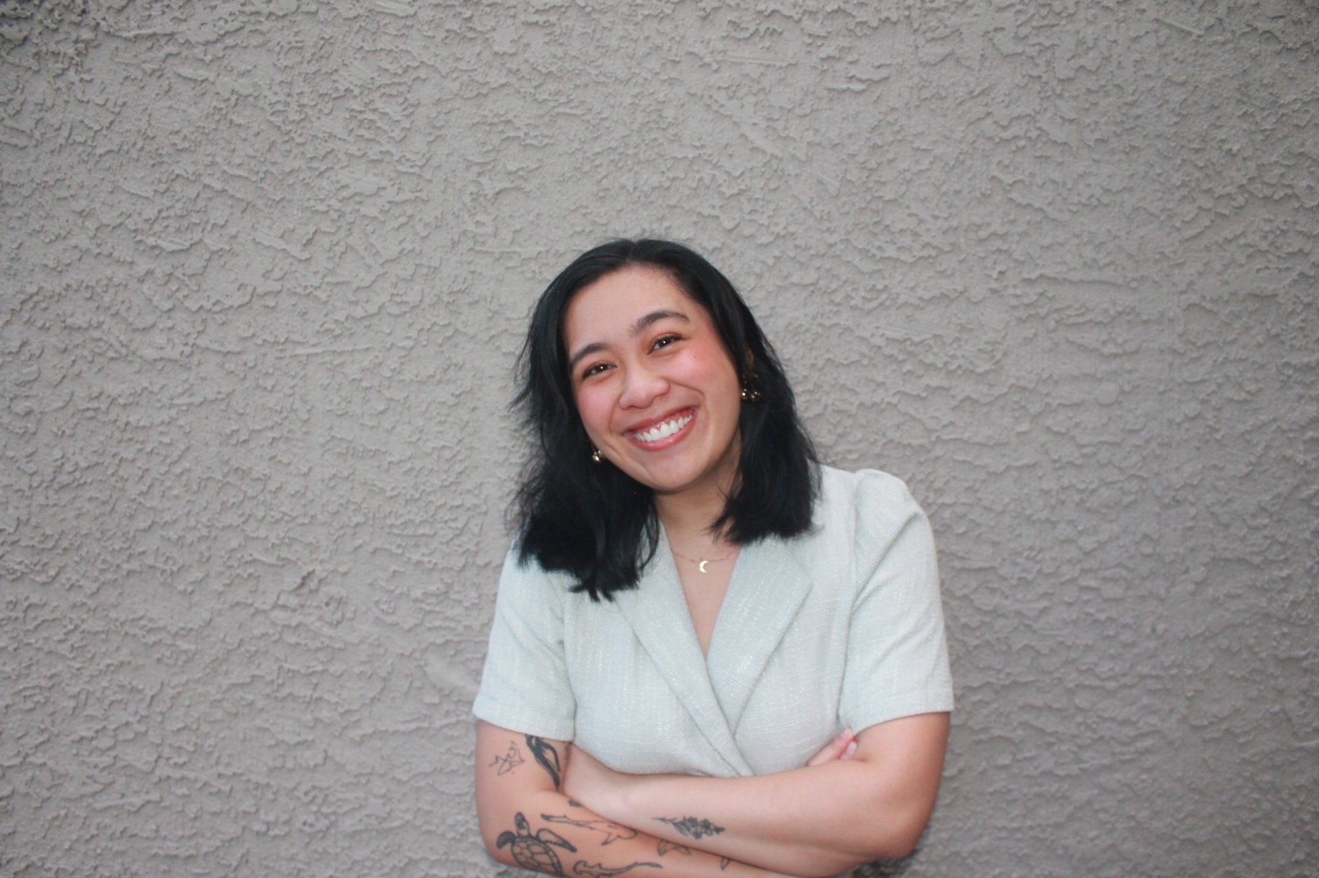 Smiling woman with black hair and arm tattoos posing against a textured grey wall.