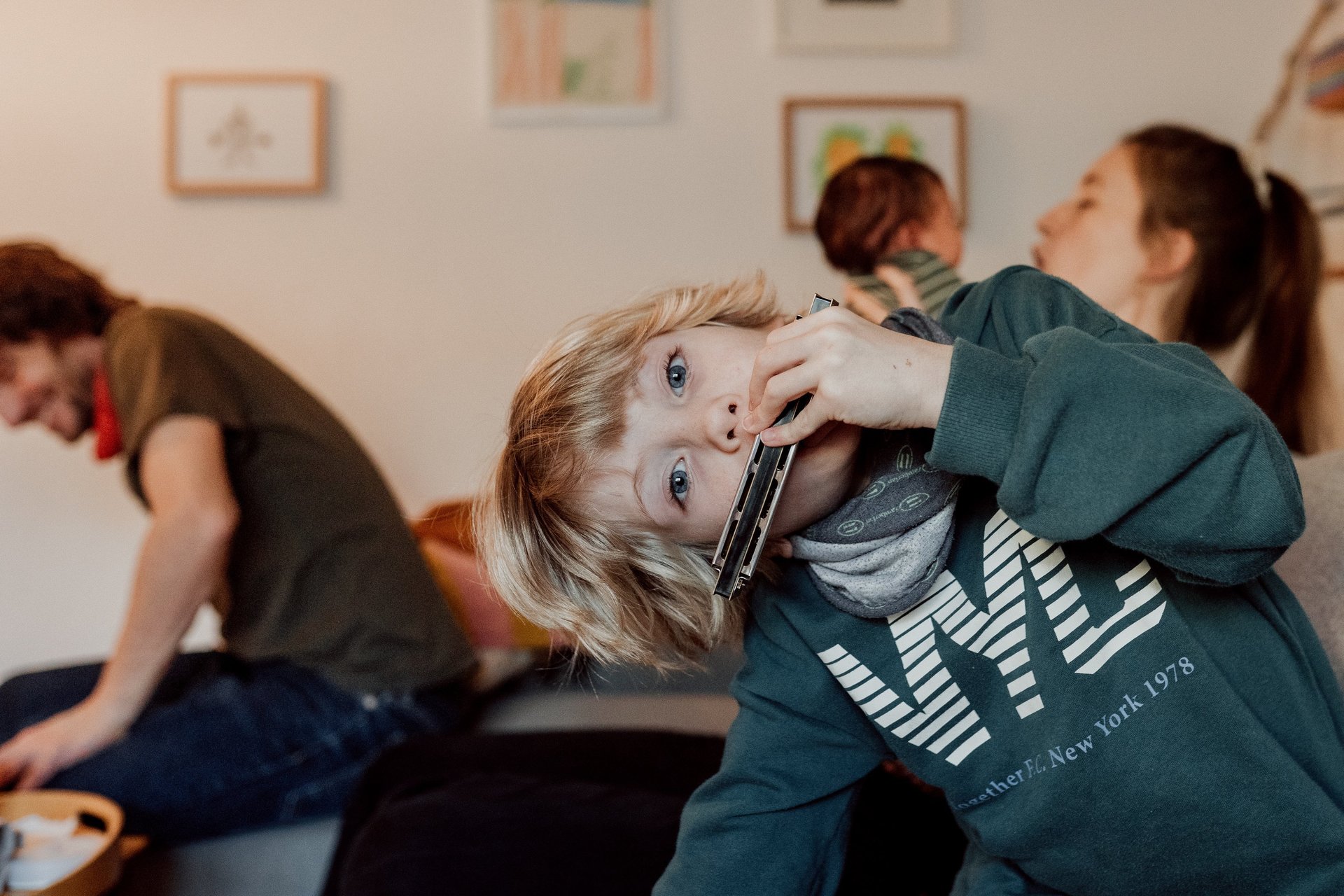 Ein junger blonder Junge spielt Mundharmonika, während sich seine Familie in einem Wohnzimmer