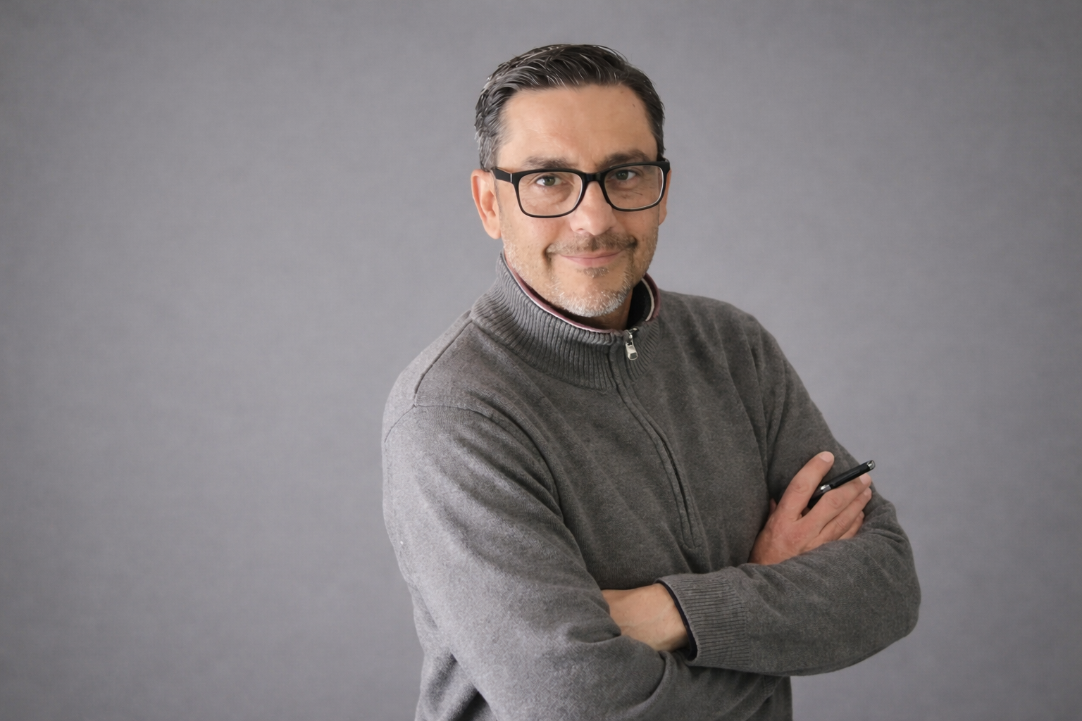 Professional portrait of a man with glasses wearing a grey quarter-zip sweater against a grey background.