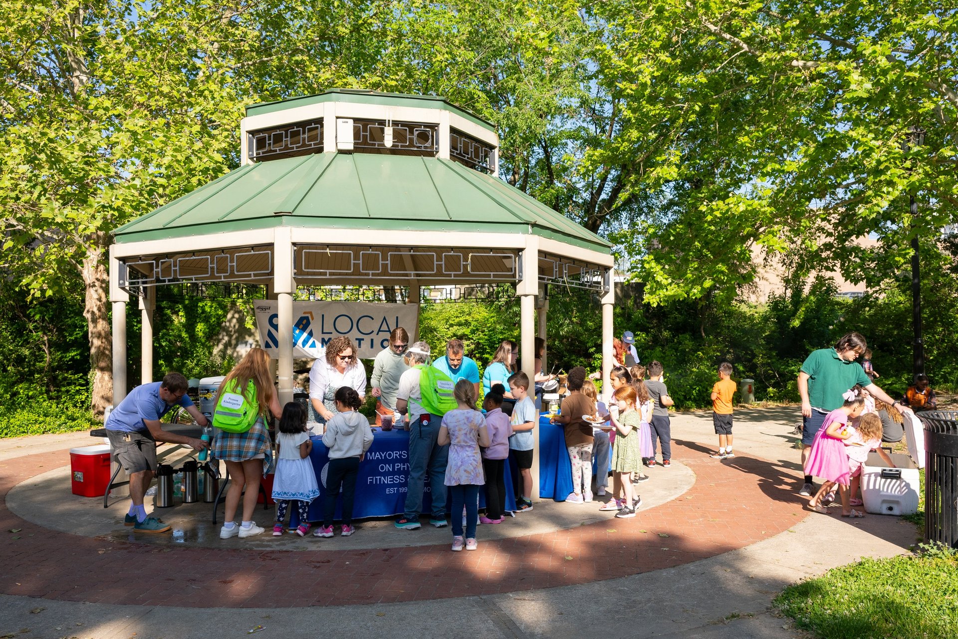 A bunch of children with a few adults approaching a table of adults in a park