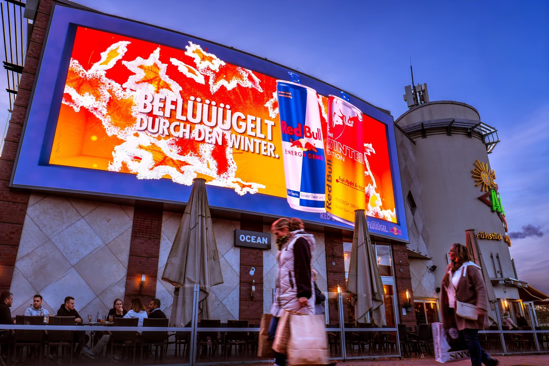Large digital billboard advertising Red Bull Winter Edition above a crowded outdoor cafe at dusk.