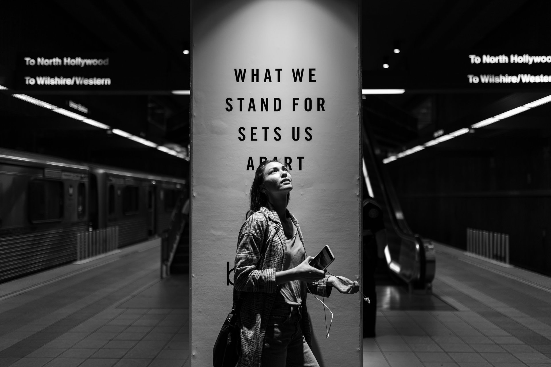 grayscale photo a woman standing in a subway station next to a social impact ad