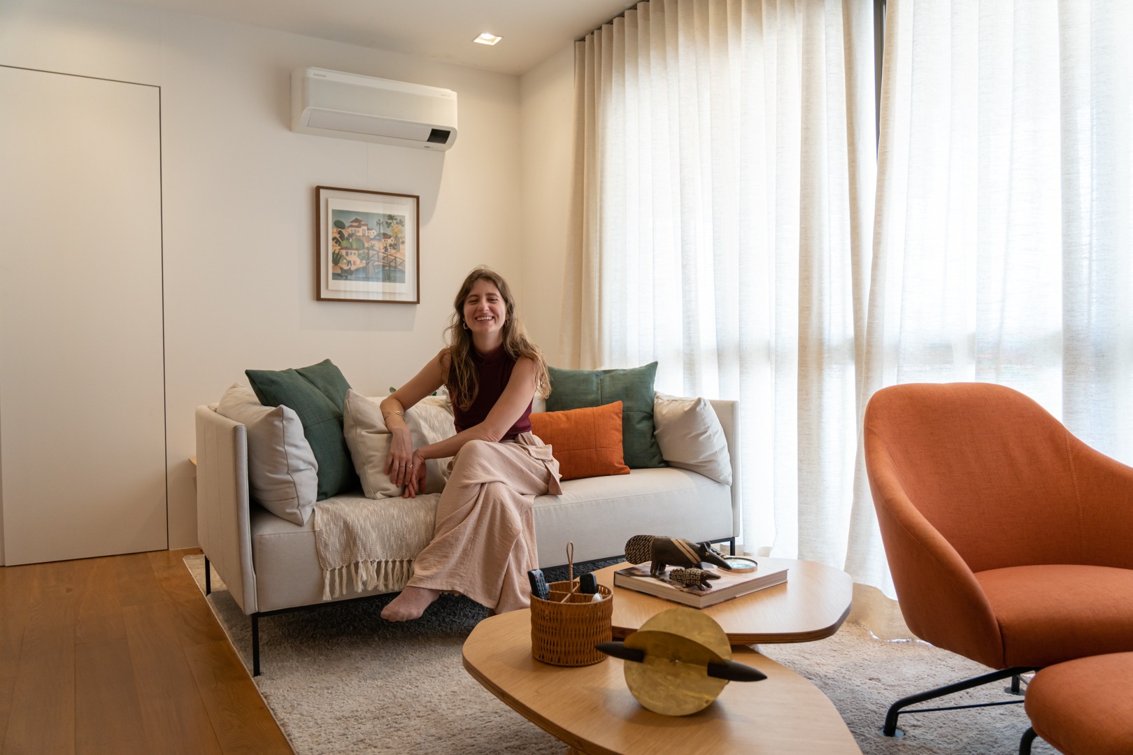 Mulher jovem sorrindo, sentada descalça em um sofá com almofadas coloridas, em uma sala aconchegante