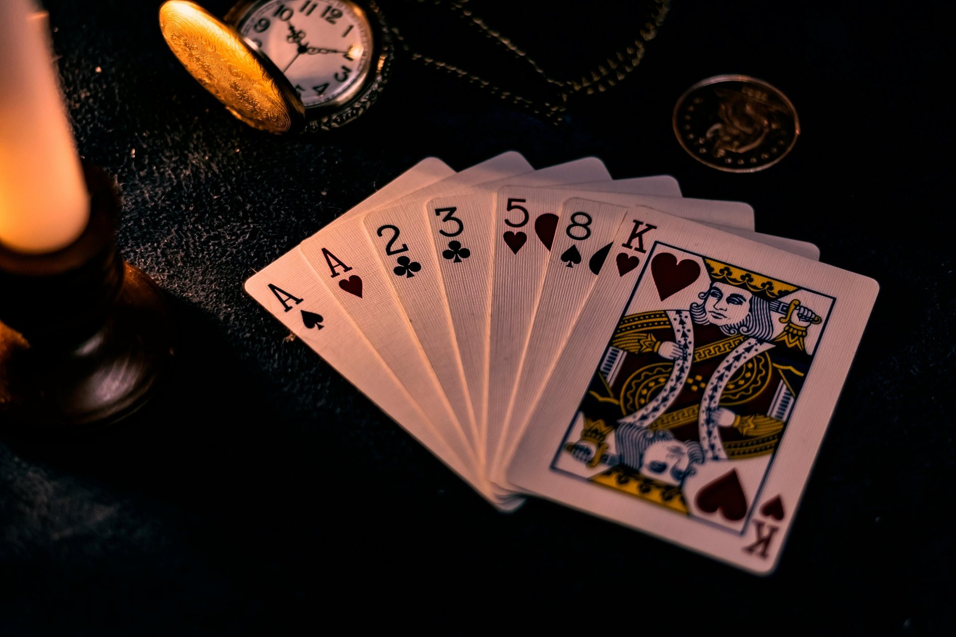 A spread hand of playing cards, witha pocketwatch, coin, chain and candlestick on the same surface in the background.
