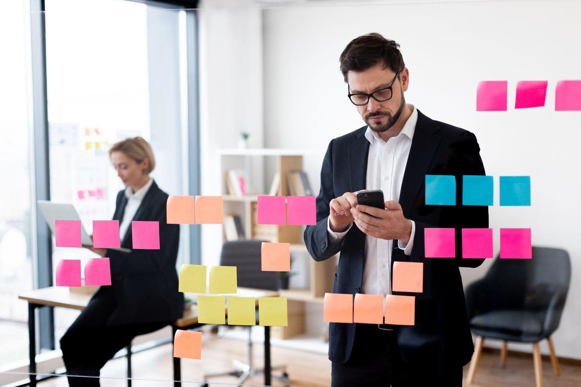 A man in a suit uses his smartphone, surrounded by colorful sticky notes on glass.