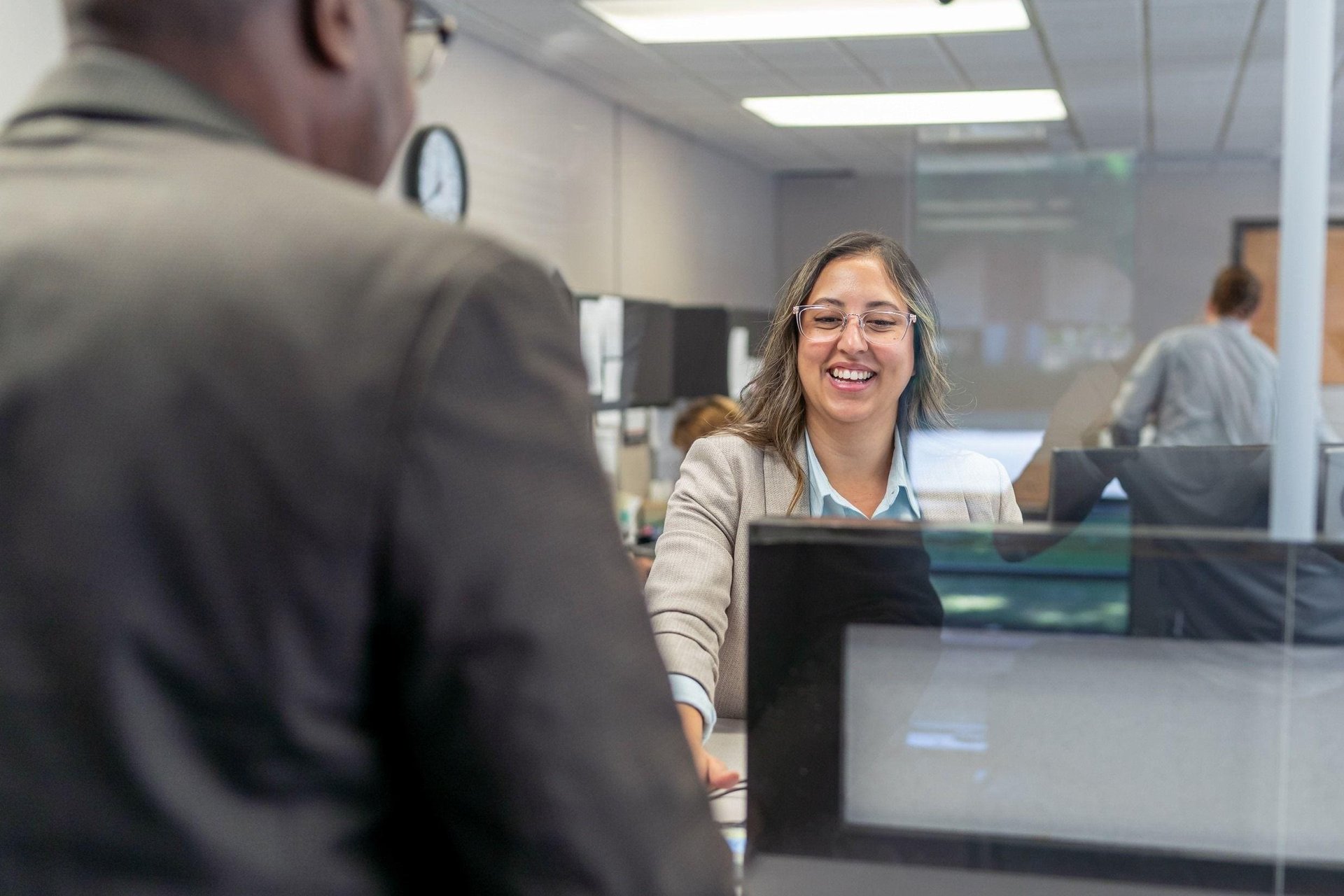 A woman in office attire smiles warmly at a customer across a reception desk.