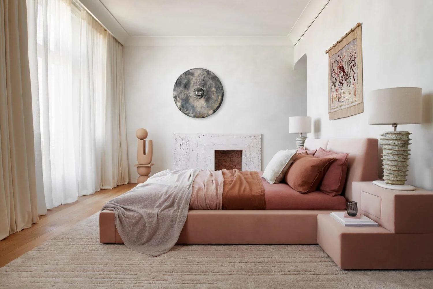 Cozy pink bedroom with layered pillows, warm bedside lighting, and neutral wall decor