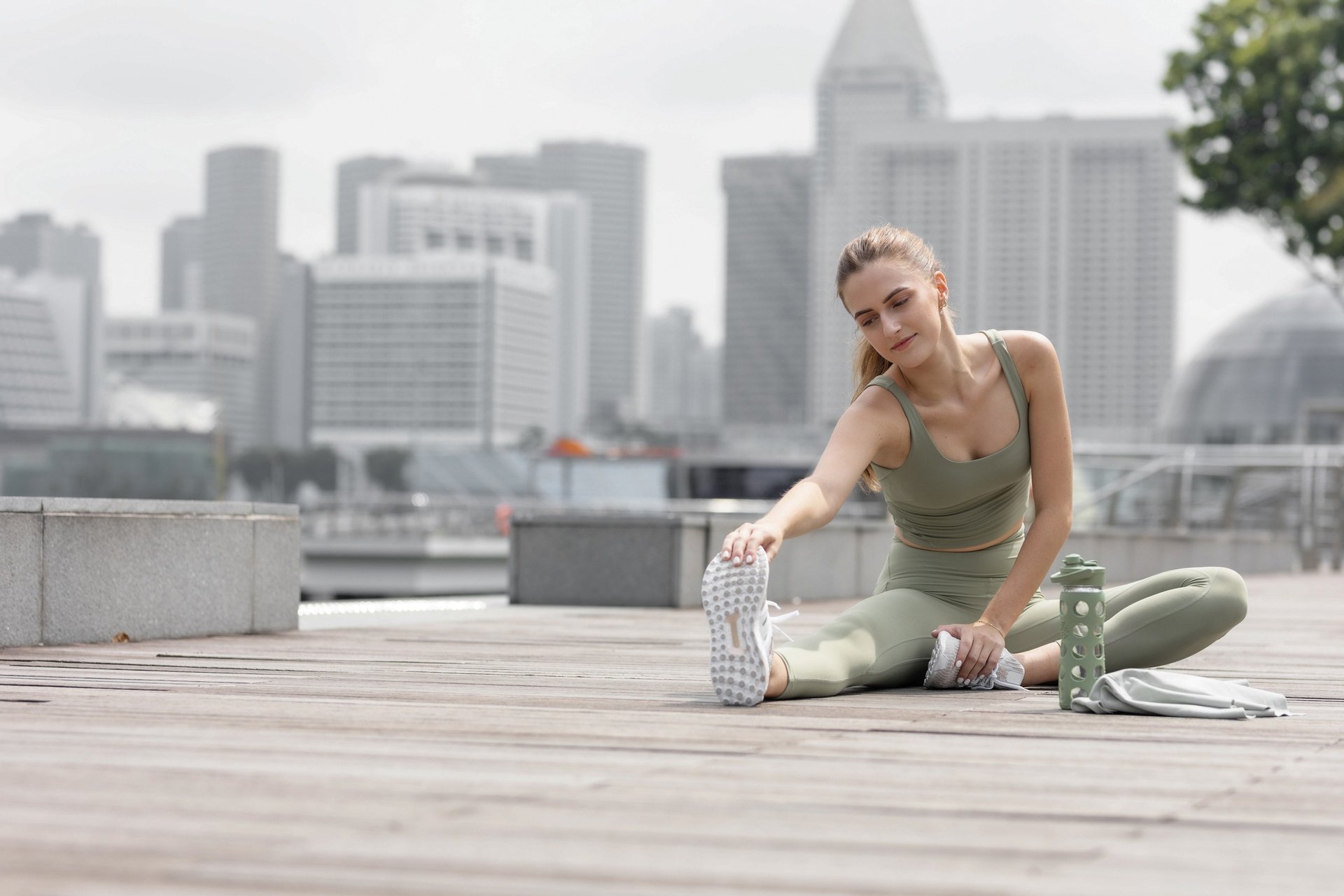 A woman wearing athletic wear, sitting outside with an urban background, stretching before exercising.
