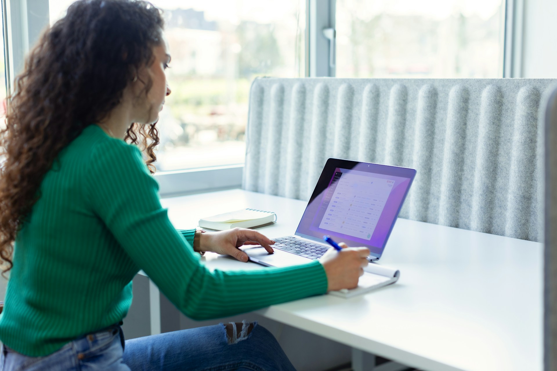 A woman with curly hair works on a laptop at a bright office desk with a notebook and pen..