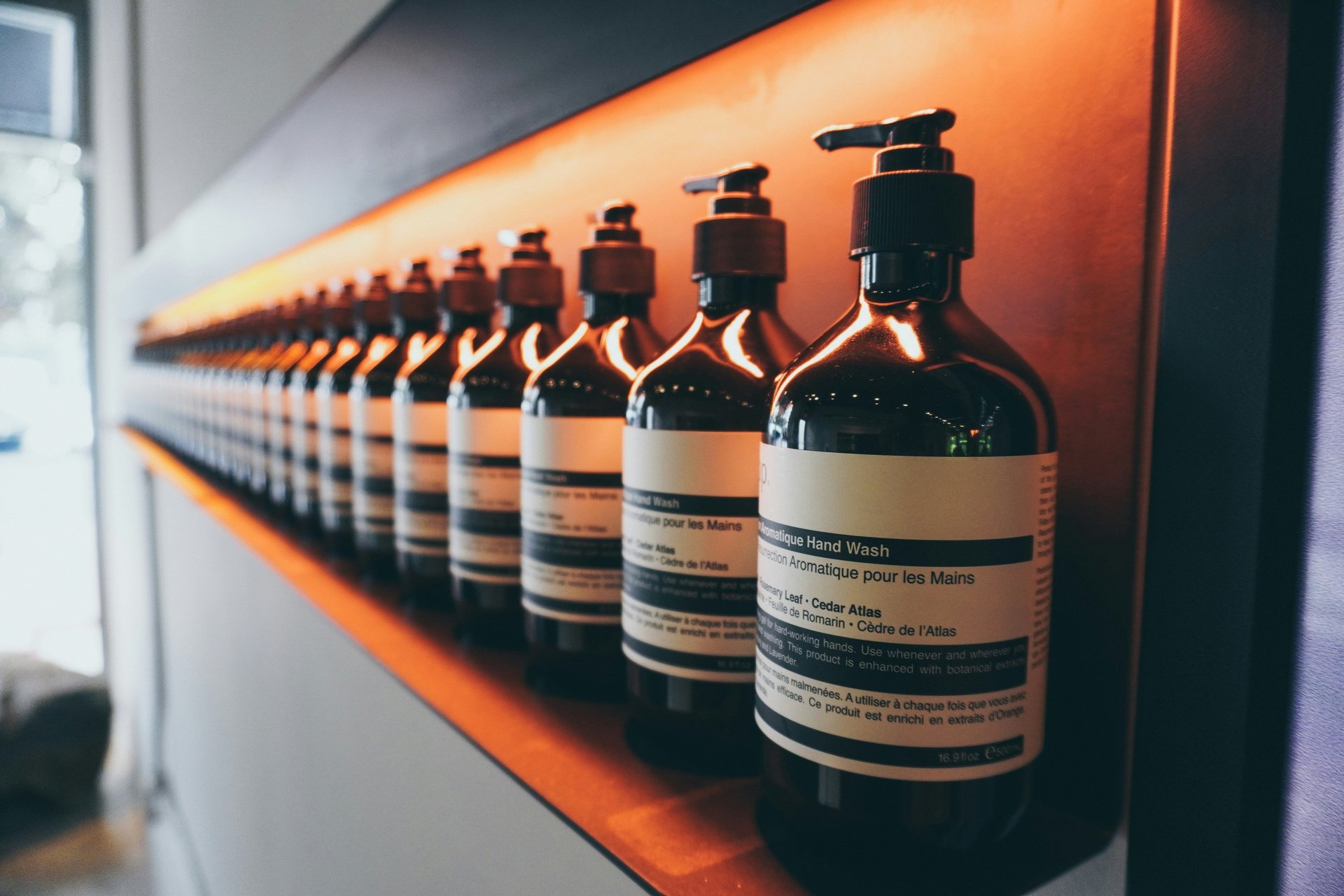 A row of Aesop aromatic hand wash glass pump bottles on a backlit wooden shelf.