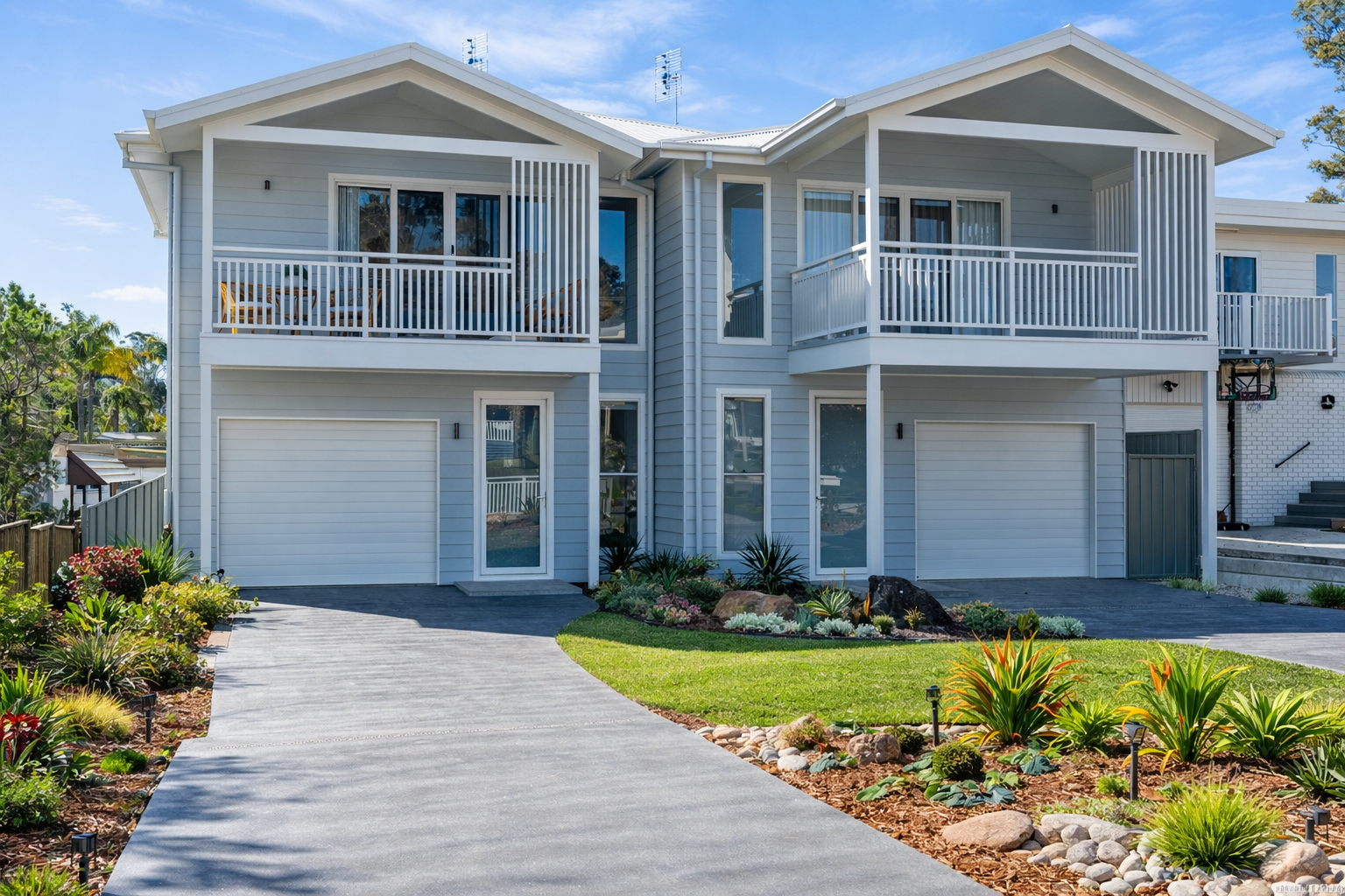 Modern two-story light blue duplex with white balconies, double garages, and a landscaped driveway.