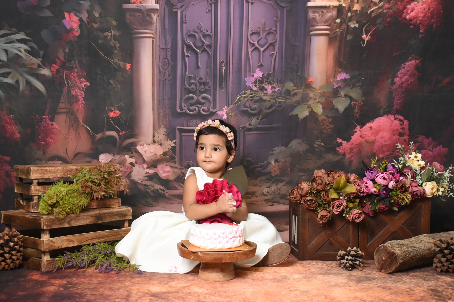 Adorable baby girl in a pink dress, celebrating her first birthday with "ONE" blocks, balloons, and floral backdrop.