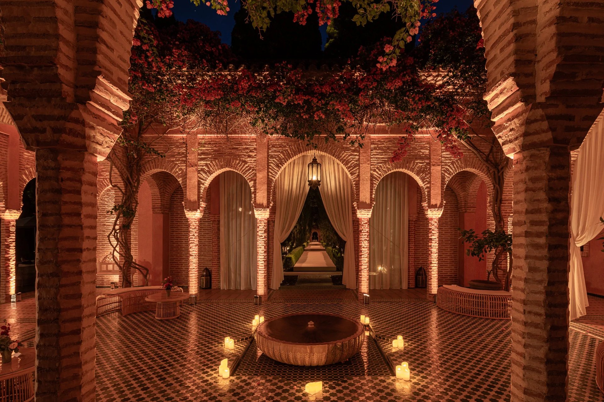 Luxury Moroccan courtyard at night with arched architecture, candlelit fountain, and pink bougainvillea.