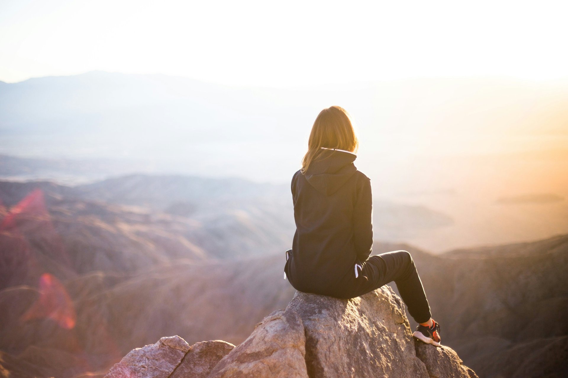 Woman sitting on mountain peak at sunrise, reflecting in solitude and enjoying peaceful nature view.