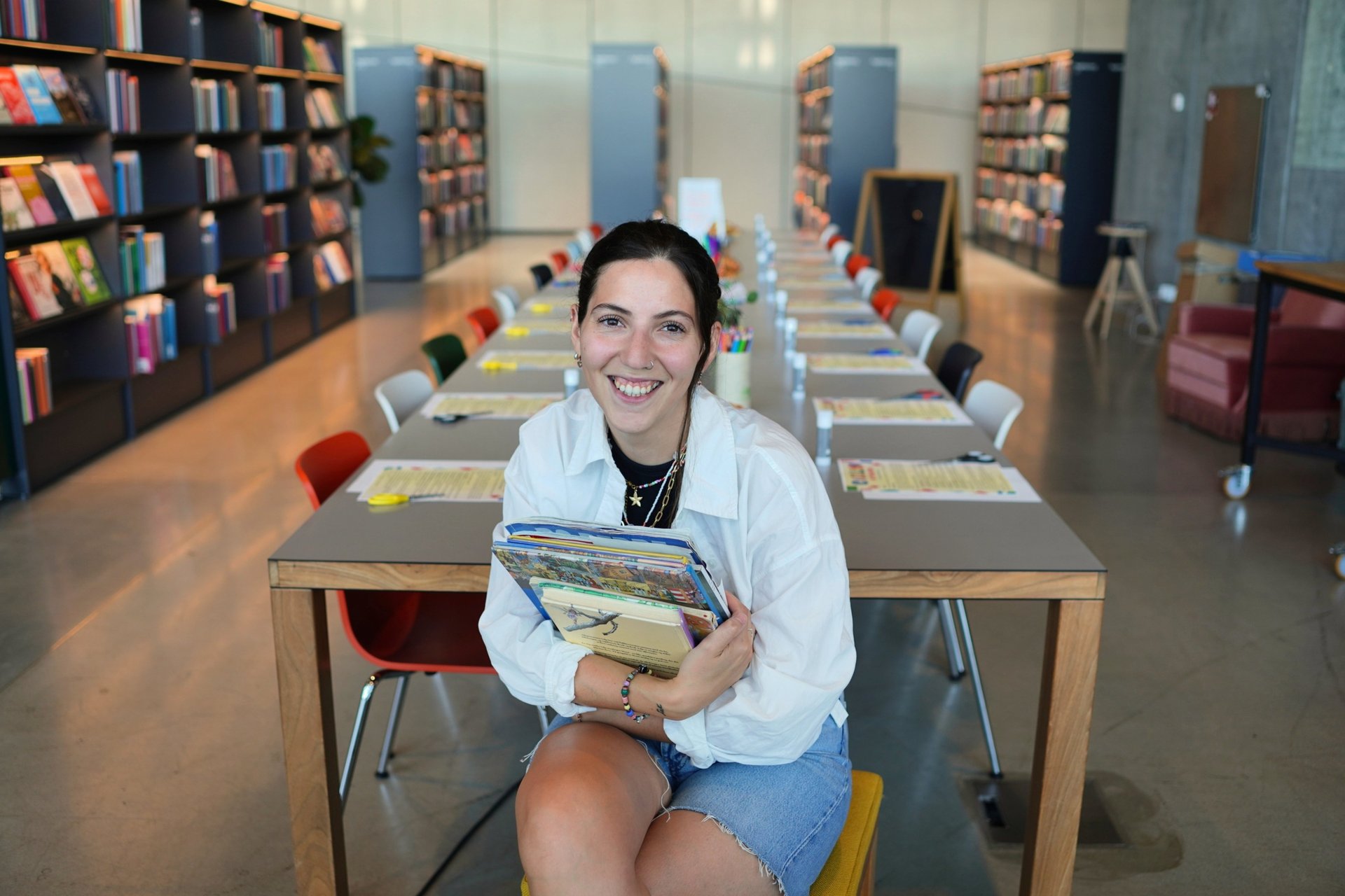 Smiling woman holding a stack of books while sitting in a modern library with study tables.