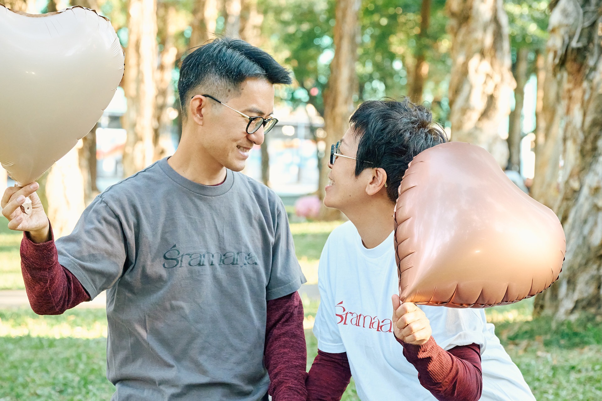 A smiling couple holds heart-shaped balloons while looking at each other in a sunny park.