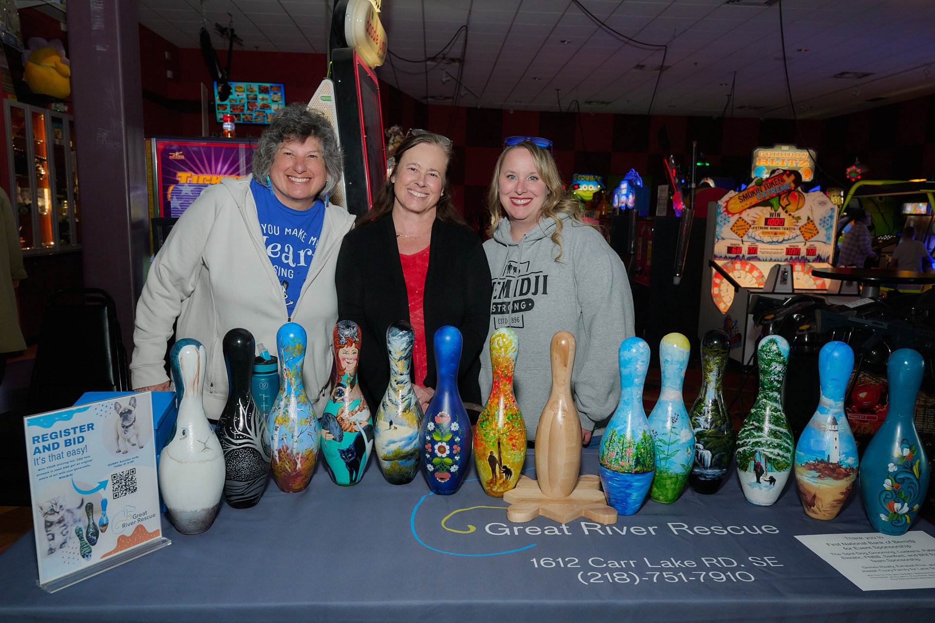 Three women smiling behind a display of hand-painted bowling pins for a Great River Rescue charity event.