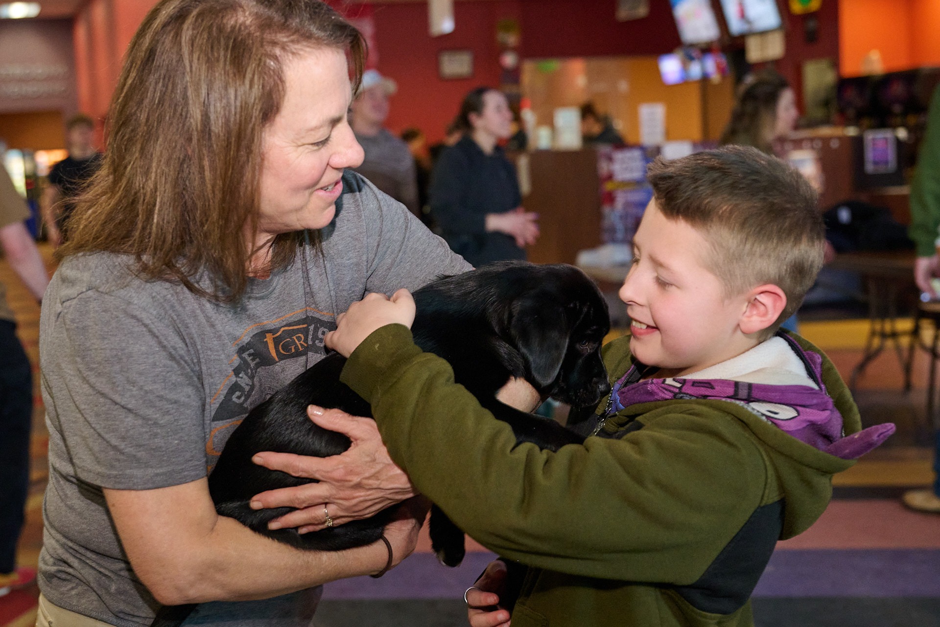 a woman holding a black puppy in her arms and a boy petting it