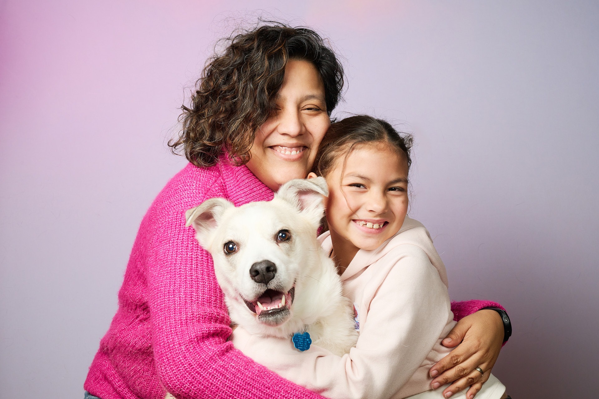 A smiling mother and daughter hugging their happy white pet dog against a purple background.