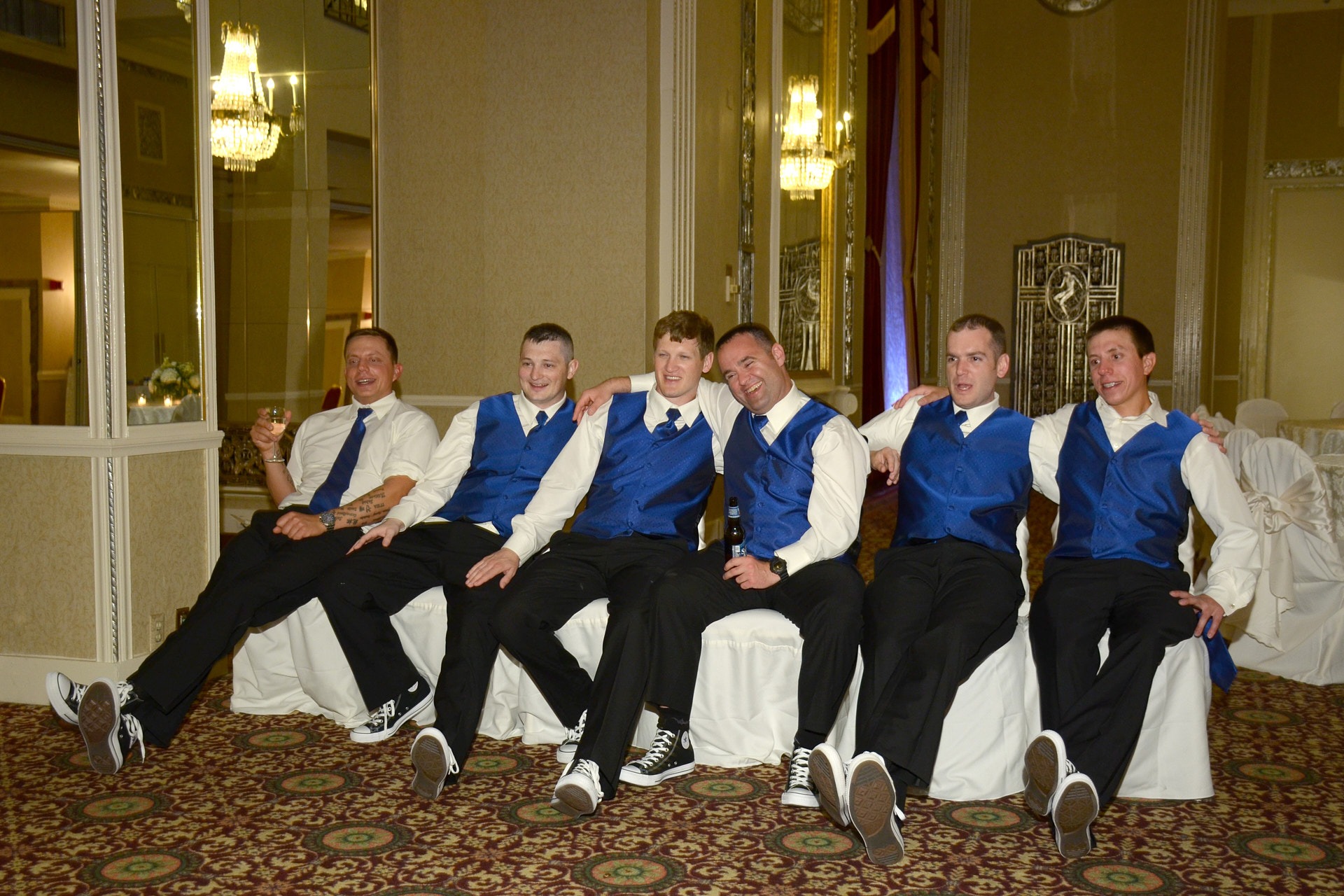 a group of men in suits and blue ties and blue vests sitting on white chairs at a hotel ball room