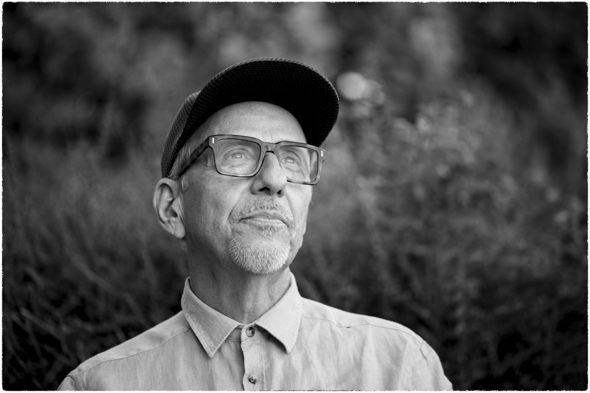 Black and white portrait of a senior man with glasses and a cap looking upwards outdoors.