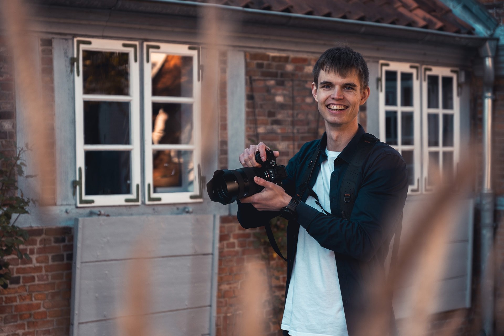 Lächelnder Hochzeitsfotograf Joel Pingel mit Kamera vor Backsteingebäude. Portrait: Just Perspectives.